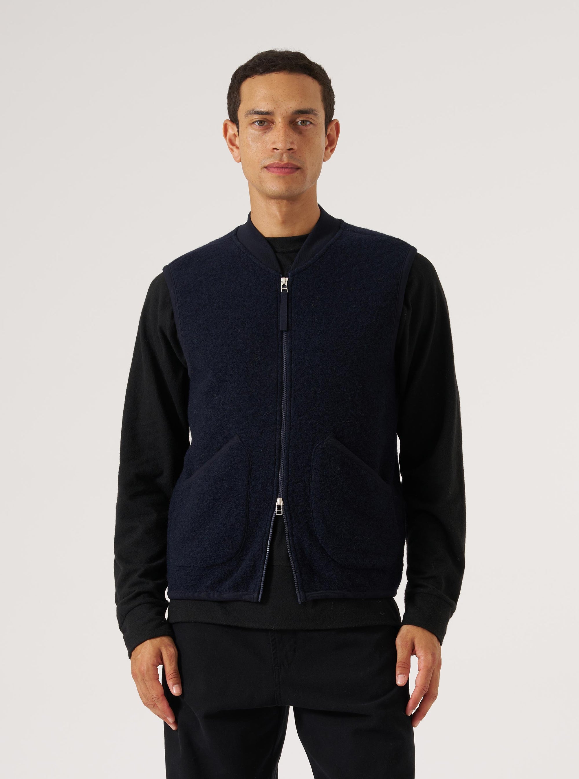 A model poses in a Navy Wool Fleece Zip Waistcoat against a white backdrop. The waistcoat is zipped up, revealing a black long-sleeve shirt underneath.