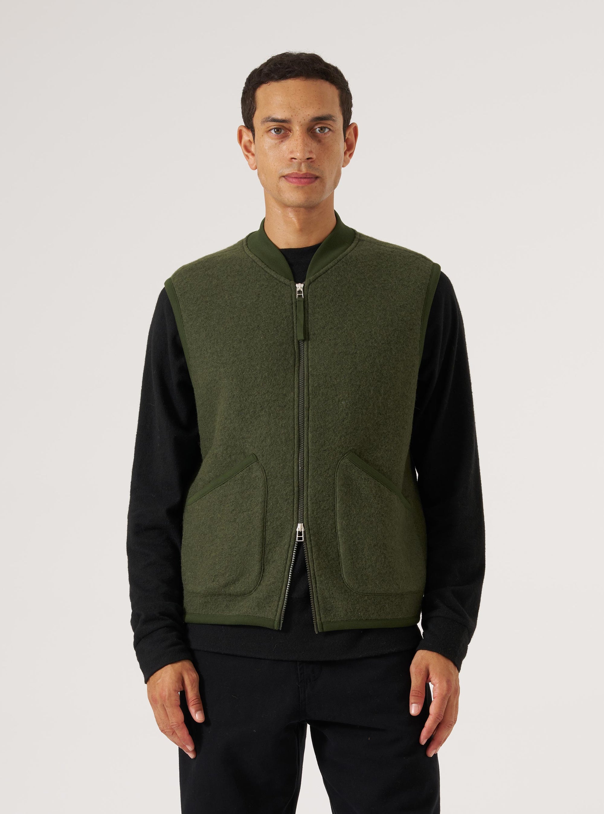 A man models the Olive Wool Fleece Zip Waistcoat against a white backdrop. The olive green waistcoat features a zip front and patch pockets.