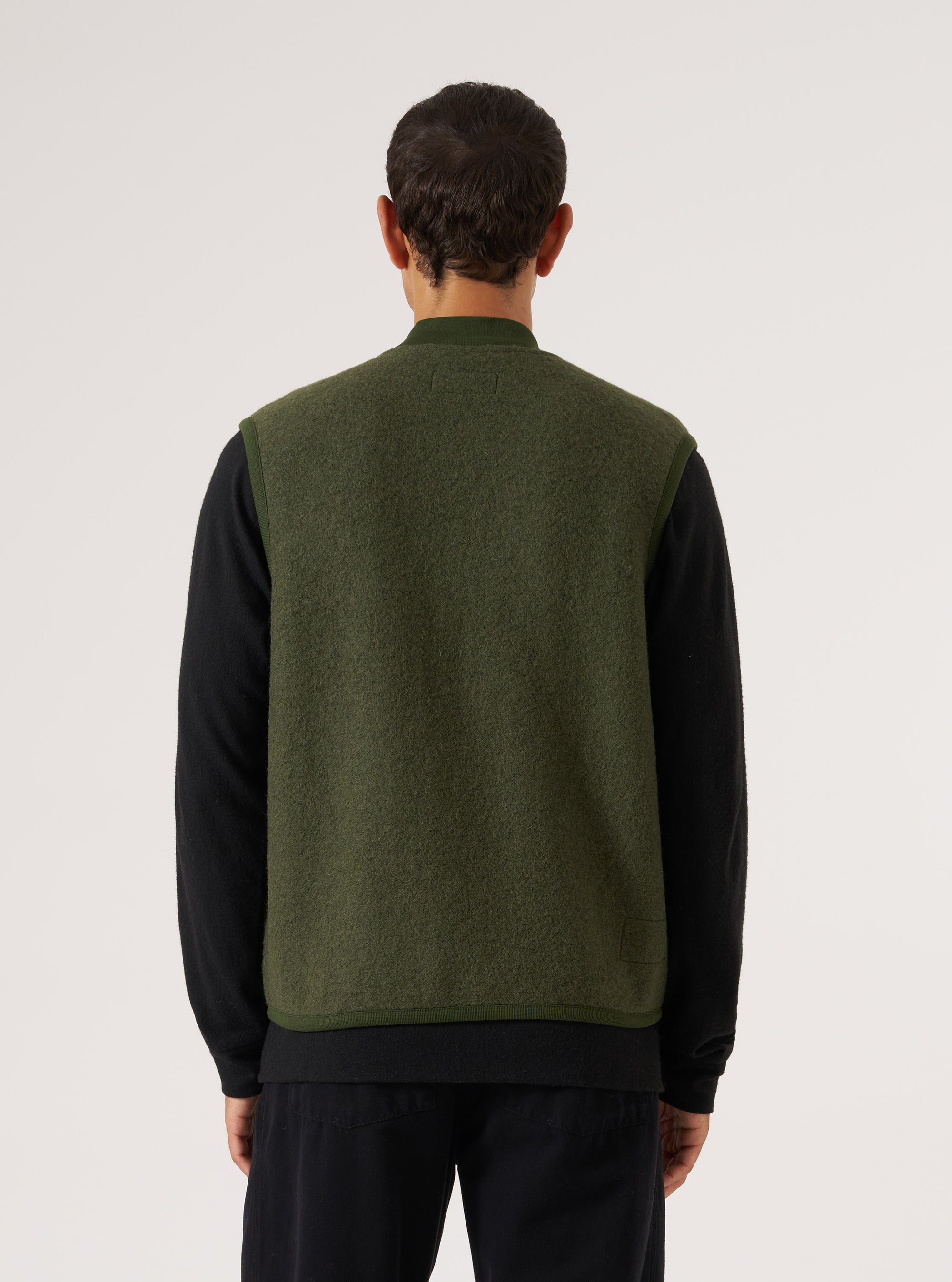 Back view of a model wearing an Olive Wool Fleece Zip Waistcoat. The olive green wool vest is layered over a black long-sleeve shirt. Shot against a white backdrop.