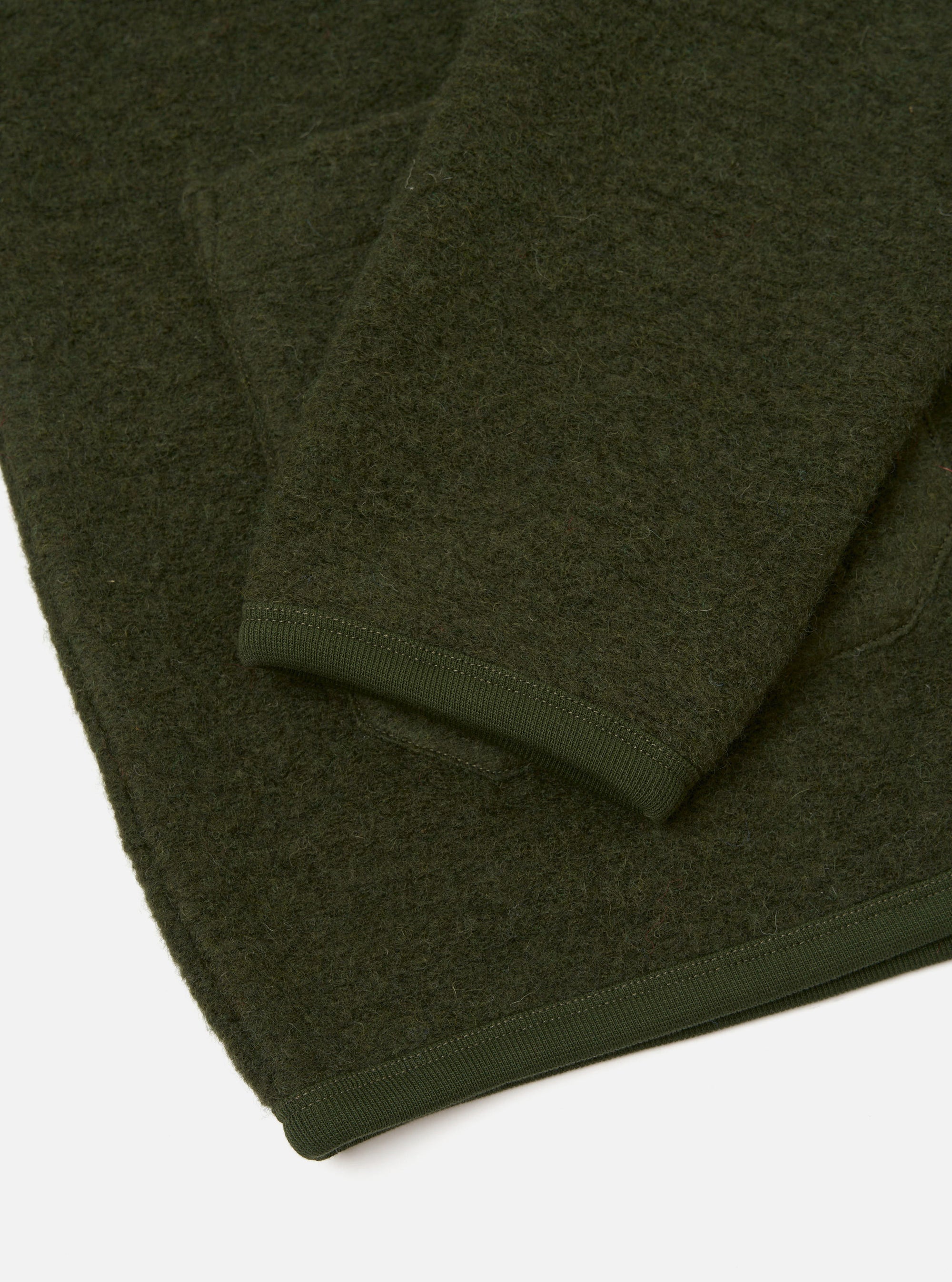 Close-up flat lay of an olive wool fleece cardigan on a white studio surface. The boiled wool texture is visible across the body and sleeve. A ribbed knit cuff in matching olive sits at the sleeve end, with the hem also finished in the same tonal ribbed trim.