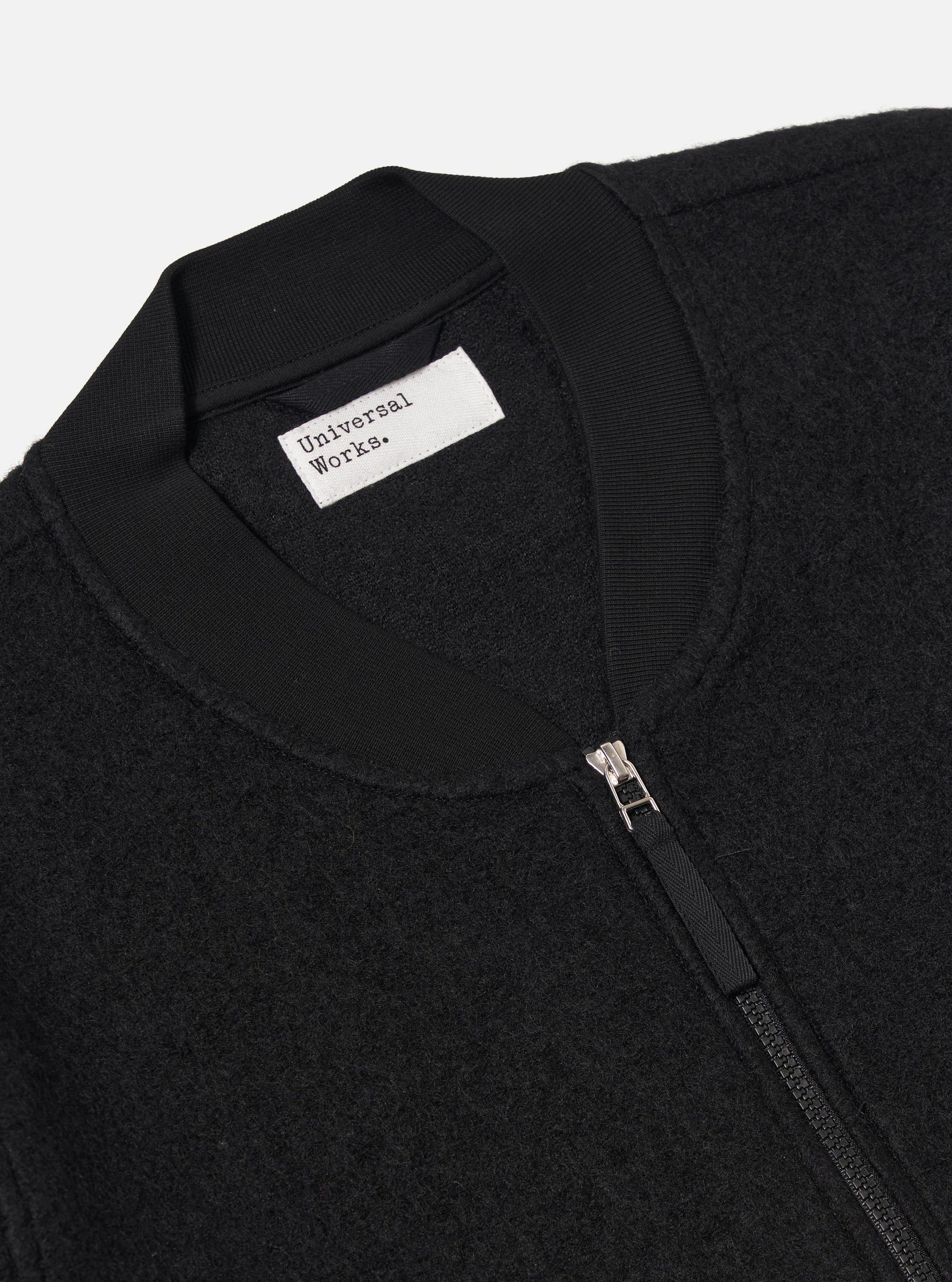 Close-up of a Black Wool Fleece Zip Bomber on a white background. The bomber has a ribbed collar and a silver zipper. The fabric is a textured wool fleece.