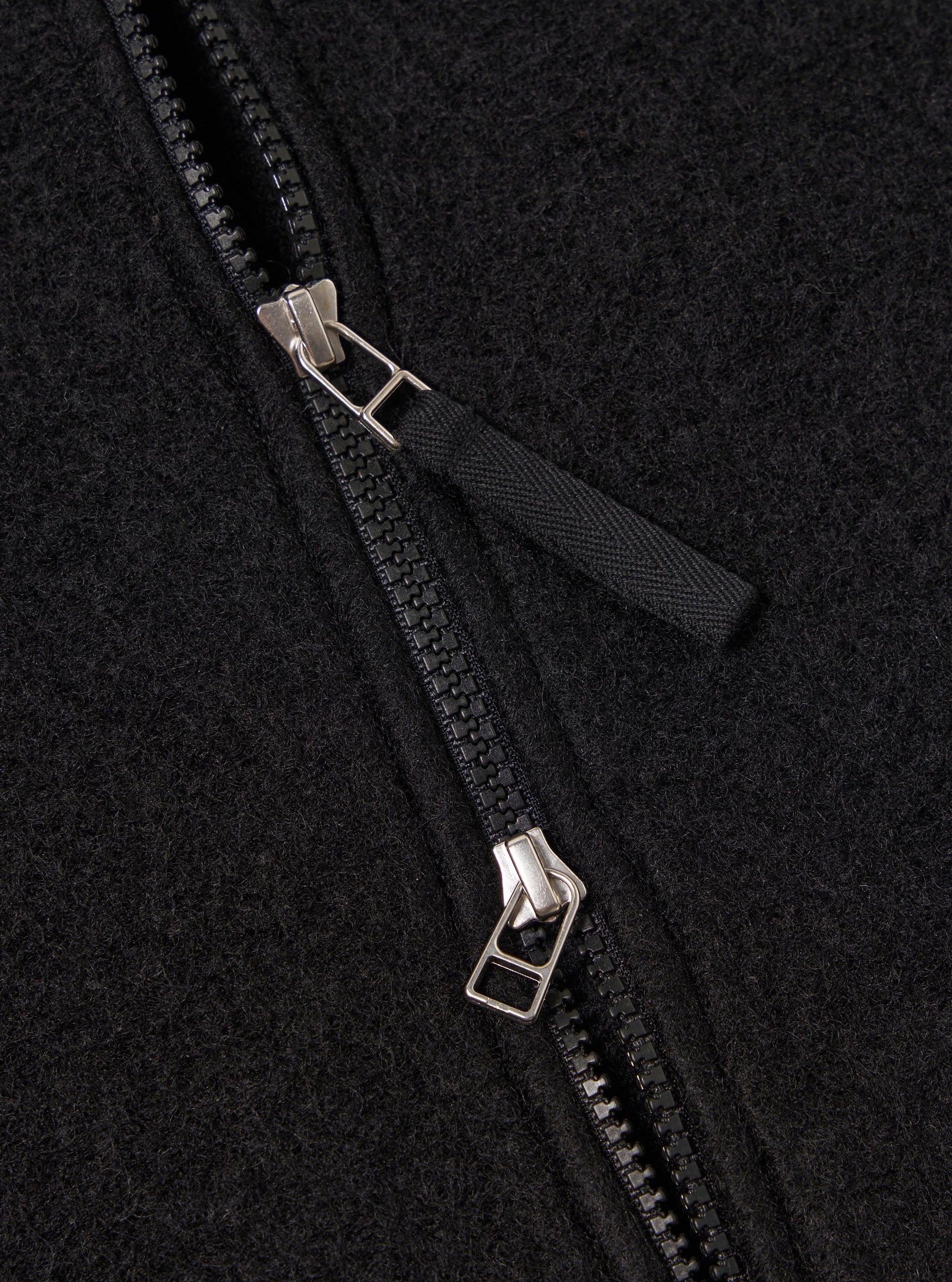 Close-up of the Black Wool Fleece Zip Bomber, showcasing the texture of the black wool fleece and the zipper detail. The background is a simple, neutral surface.