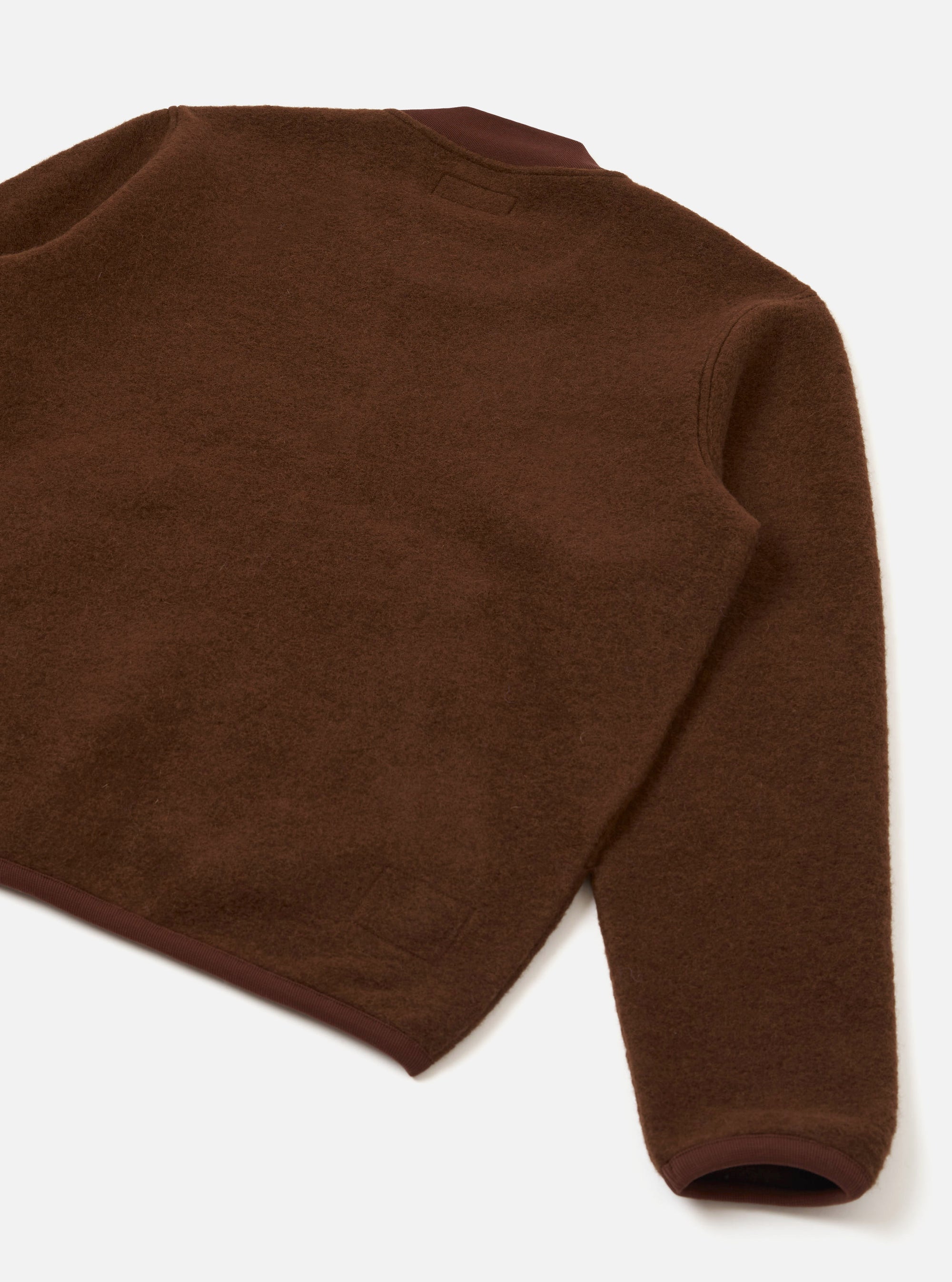 Flat lay of a cacao wool fleece zip bomber on a white studio surface, shot from above at an angle. The back and one sleeve are visible, showing a dense, brushed wool fleece texture. Darker brown woven binding trims the hem and cuff. A small woven label sits at the centre back neckline.