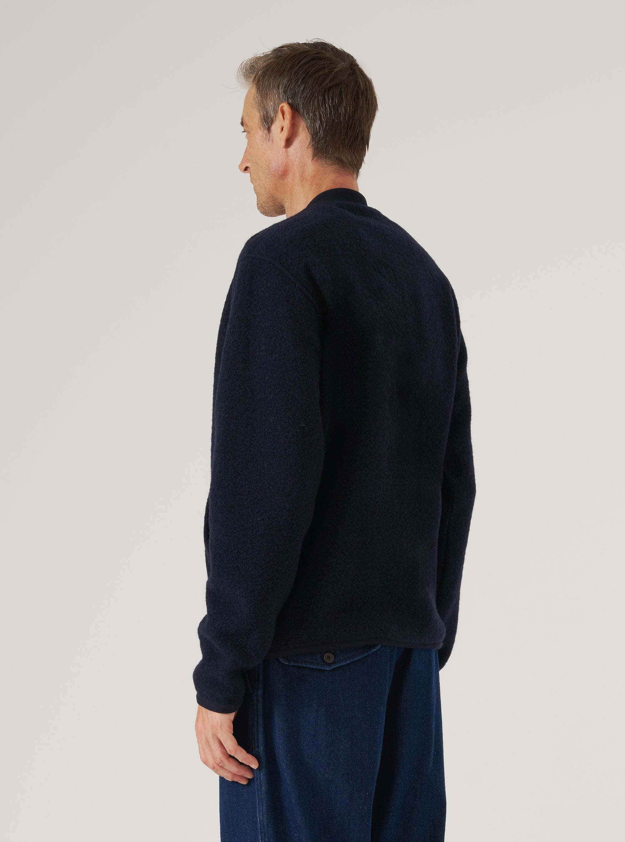 Navy wool fleece zip bomber worn by a male model, shown from a three-quarter rear angle against a light grey studio backdrop. The sweater-weight fleece has a textured, brushed surface in navy. The ribbed cuffs are visible at the wrists. The model wears it over dark indigo wide-leg trousers. Shot cropped from head to mid-thigh.