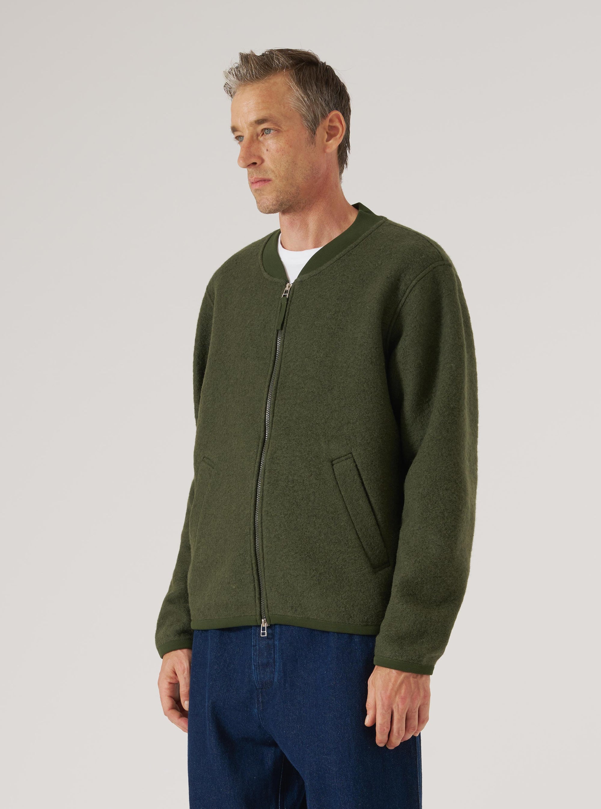 Olive wool fleece zip bomber worn by a male model on a grey studio backdrop. The jacket features a ribbed bomber collar, full-length silver zip, welt hand pockets, and ribbed cuffs. The textured boiled wool fleece is visible throughout. Worn zipped up over a white crew-neck tee, paired with dark blue jeans. Shot cropped at the thigh, three-quarter angle.