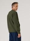 Male model photographed from behind and to the side against a light grey studio backdrop, wearing an olive wool fleece zip bomber with a ribbed crew neck collar and long sleeves with ribbed cuffs. The brushed wool texture is visible across the back. Worn untucked over dark navy trousers. Shot crops just below the hips.