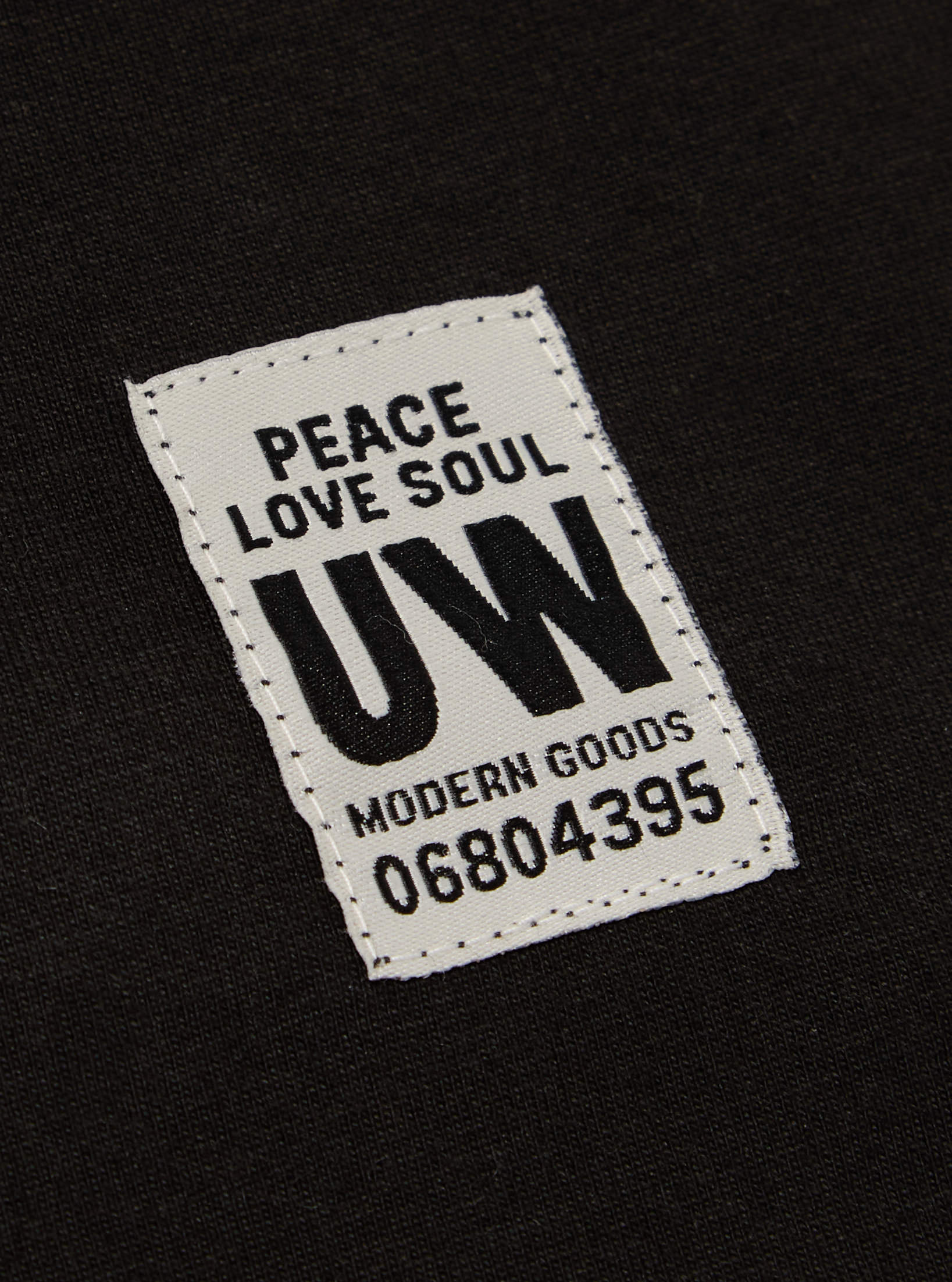 Close-up of the Black Single Jersey Core Tee showing the fabric and a tag with 'Peace Love Soul UW Modern Goods 06804395' on it. The tee is laid flat.