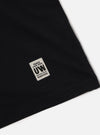Close-up of a Black Single Jersey Core Tee on a white surface, showing the fabric detail and a branded tag with 'Peace Love Soul UW' and a product code.