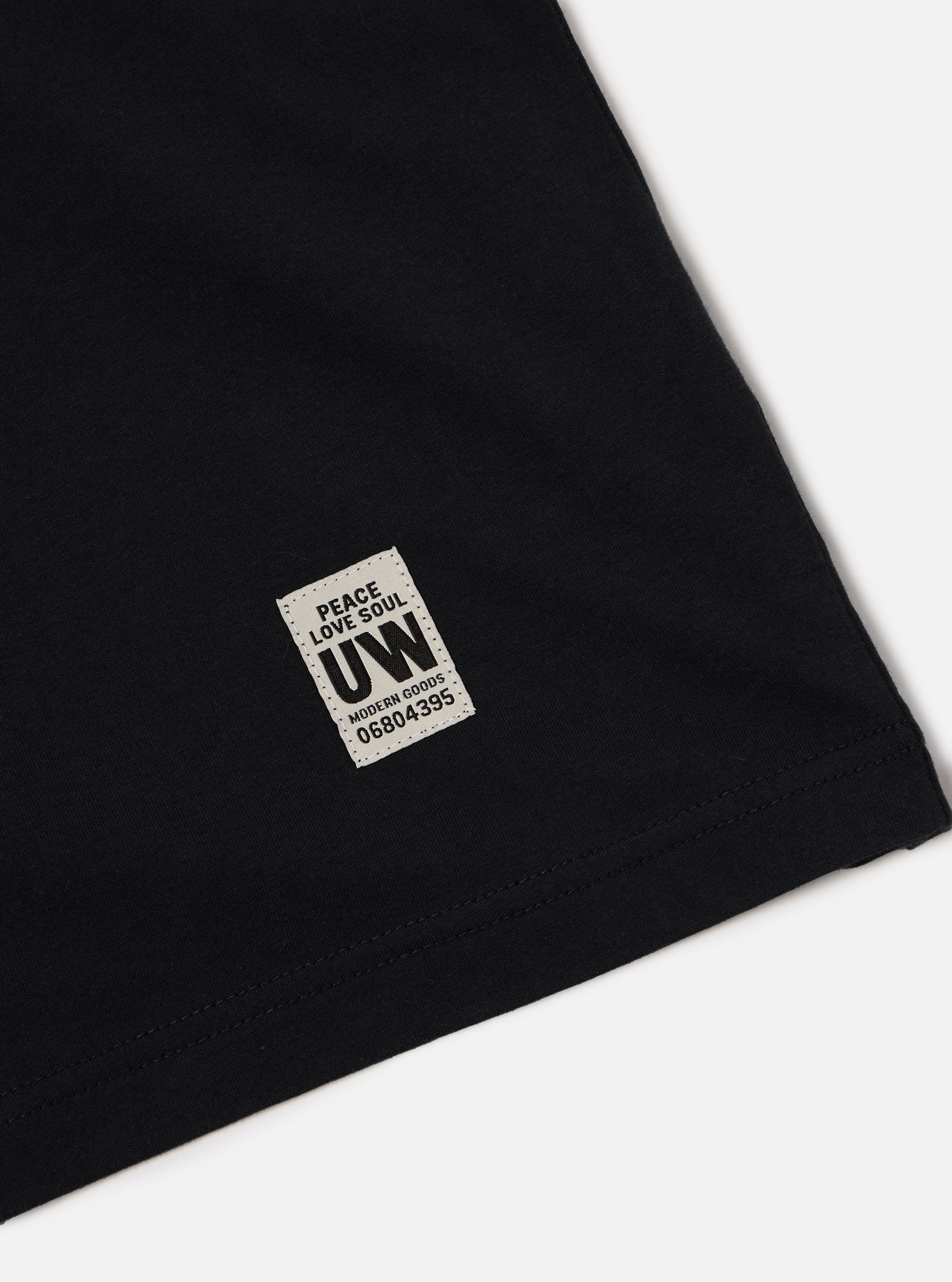 Close-up of a Black Single Jersey Core Tee on a white surface, showing the fabric detail and a branded tag with 'Peace Love Soul UW' and a product code.