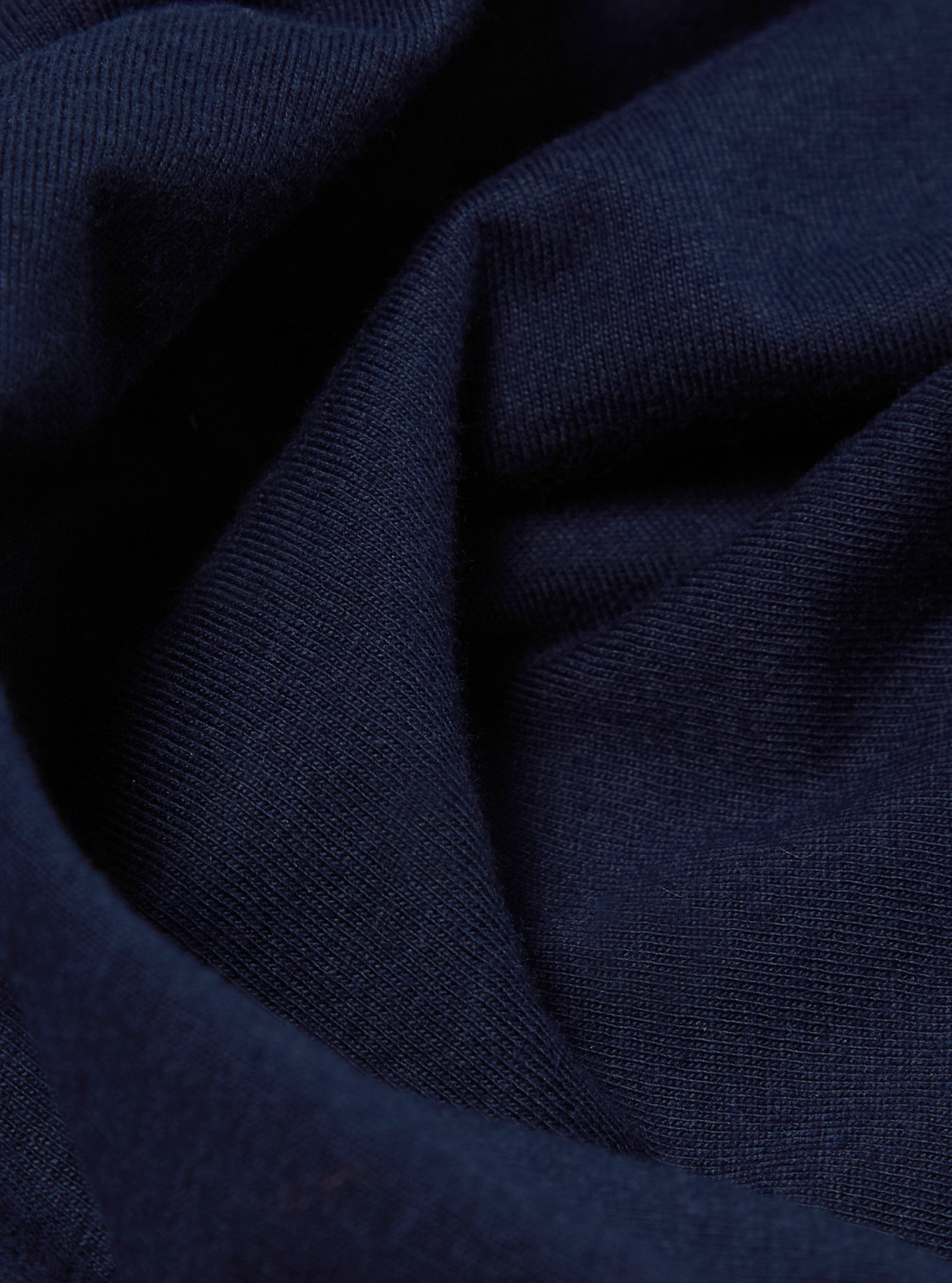 Close-up of the Navy Single Jersey Core Tee fabric. The soft, textured material is draped on a flat surface, showcasing its rich navy color and fine knit.
