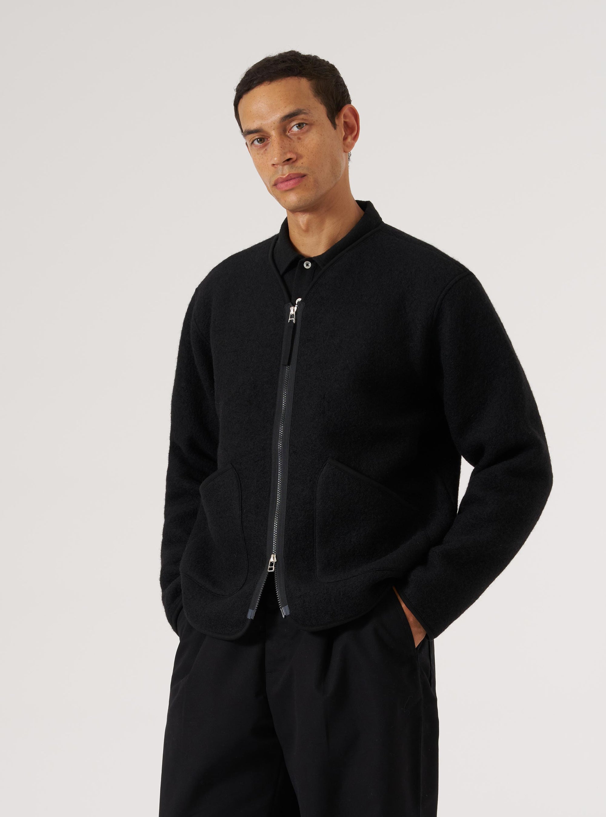 A model poses in a Black Wool Fleece Zip Liner Jacket against a white backdrop. The jacket is black wool fleece with a zip front and pockets.