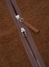 Close-up detail of the double zip fastening on a cacao wool fleece liner jacket. Two silver-hardware zip sliders sit on a dark plum zip tape with moulded plastic teeth. A woven brown zip garage tab is visible. The brushed, textured wool fleece fabric fills the background.