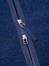 Close-up detail of the centre-front zip on an indigo wool fleece liner jacket. Two silver metal zip pullers sit on a navy zip tape with plastic teeth, partially open to reveal the garment's interior. The brushed wool fleece fabric shows a dense, textured surface in deep indigo blue.