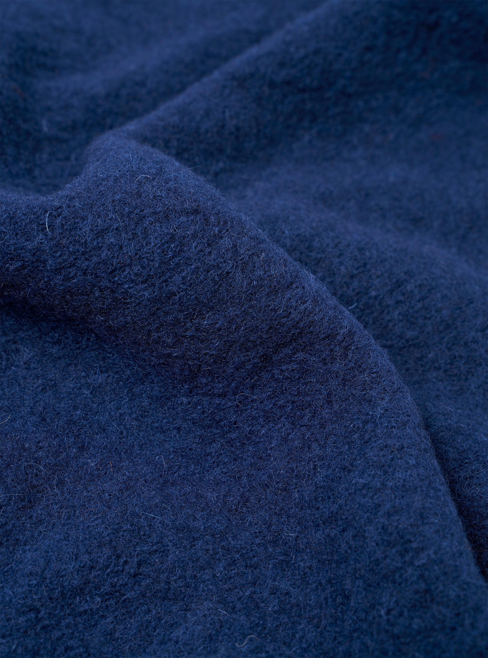 Close-up of the indigo wool fleece fabric used in the zip liner jacket. The boiled wool texture is dense and lightly brushed, with short curling fibres creating a soft, napped surface. The deep navy-indigo tone shows subtle variation across the draped folds of the cloth.