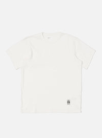 Flat lay of an Ecru Single Jersey Core Tee on a white background. The tee is a light cream color and has a relaxed fit. A small tag is visible near the hem.