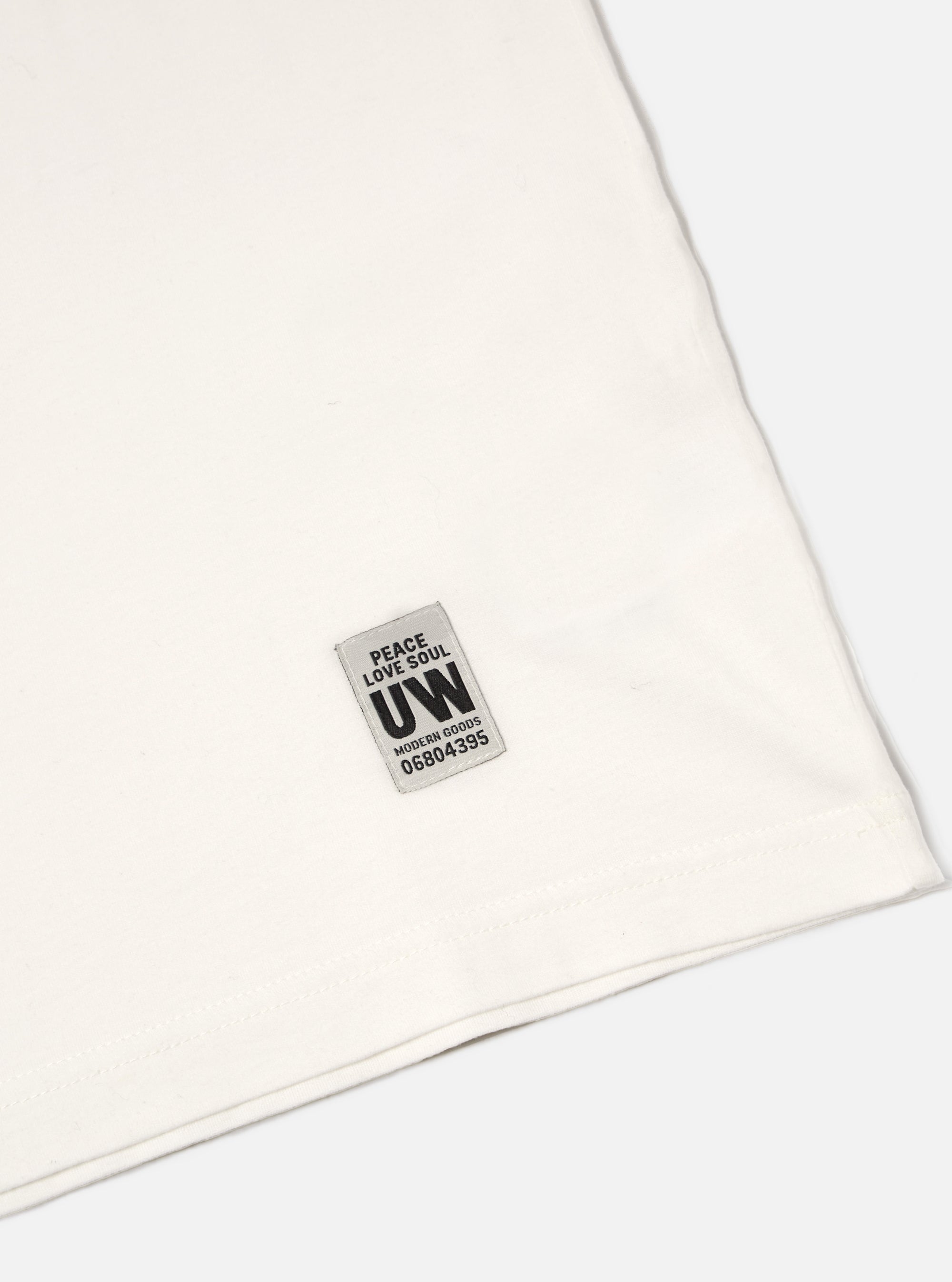 Close-up of the Ecru Single Jersey Core Tee on a white surface, showing the fabric texture and UW label. The tee has a classic, comfortable fit.