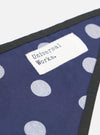 Close-up of the Navy Bold Dot Print Rayon Scout Scarf on a white surface. The scarf features a navy background with white polka dots and a black trim. A white tag with "Universal Works." is visible.