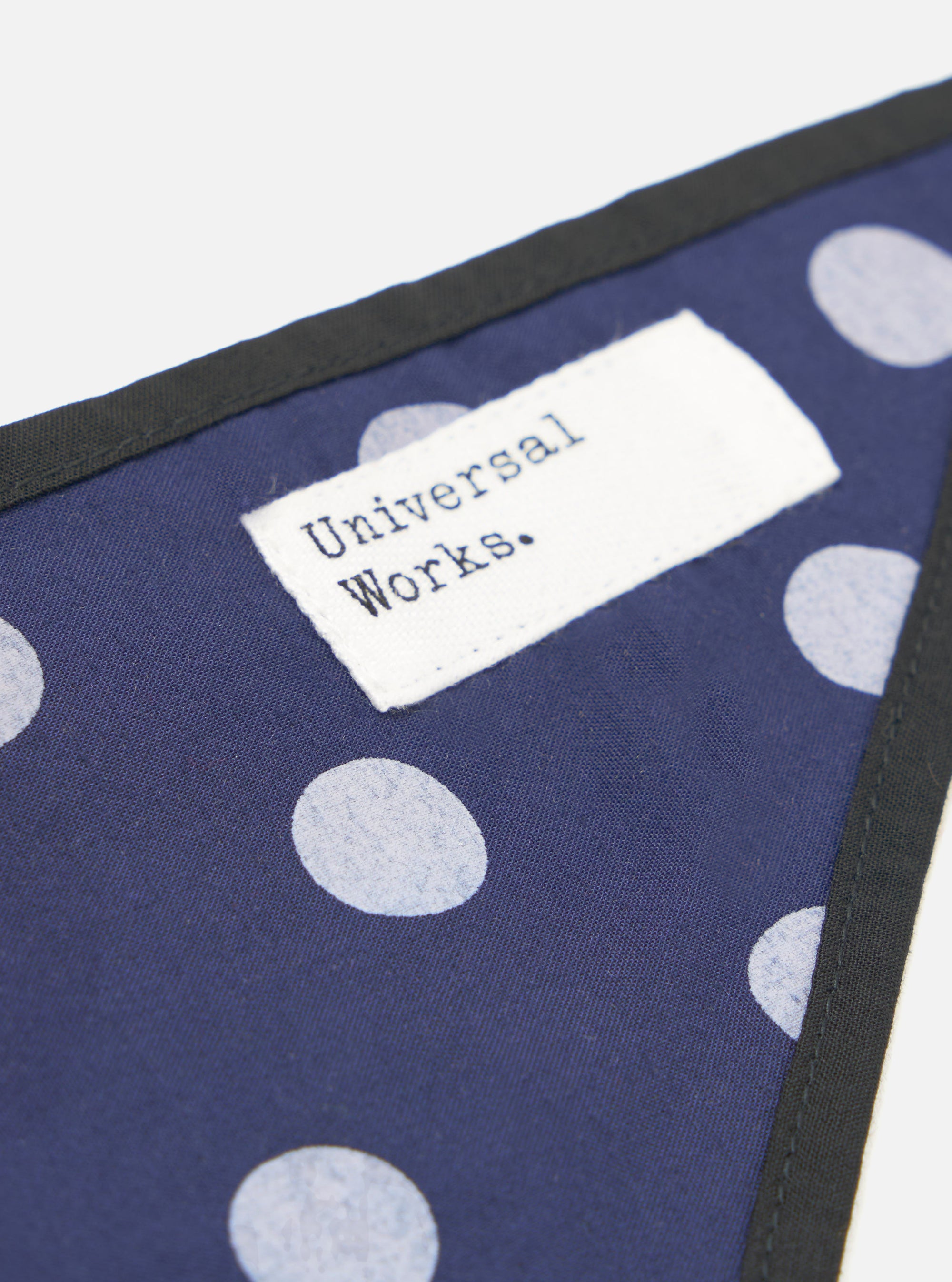 Close-up of the Navy Bold Dot Print Rayon Scout Scarf on a white surface. The scarf features a navy background with white polka dots and a black trim. A white tag with "Universal Works." is visible.
