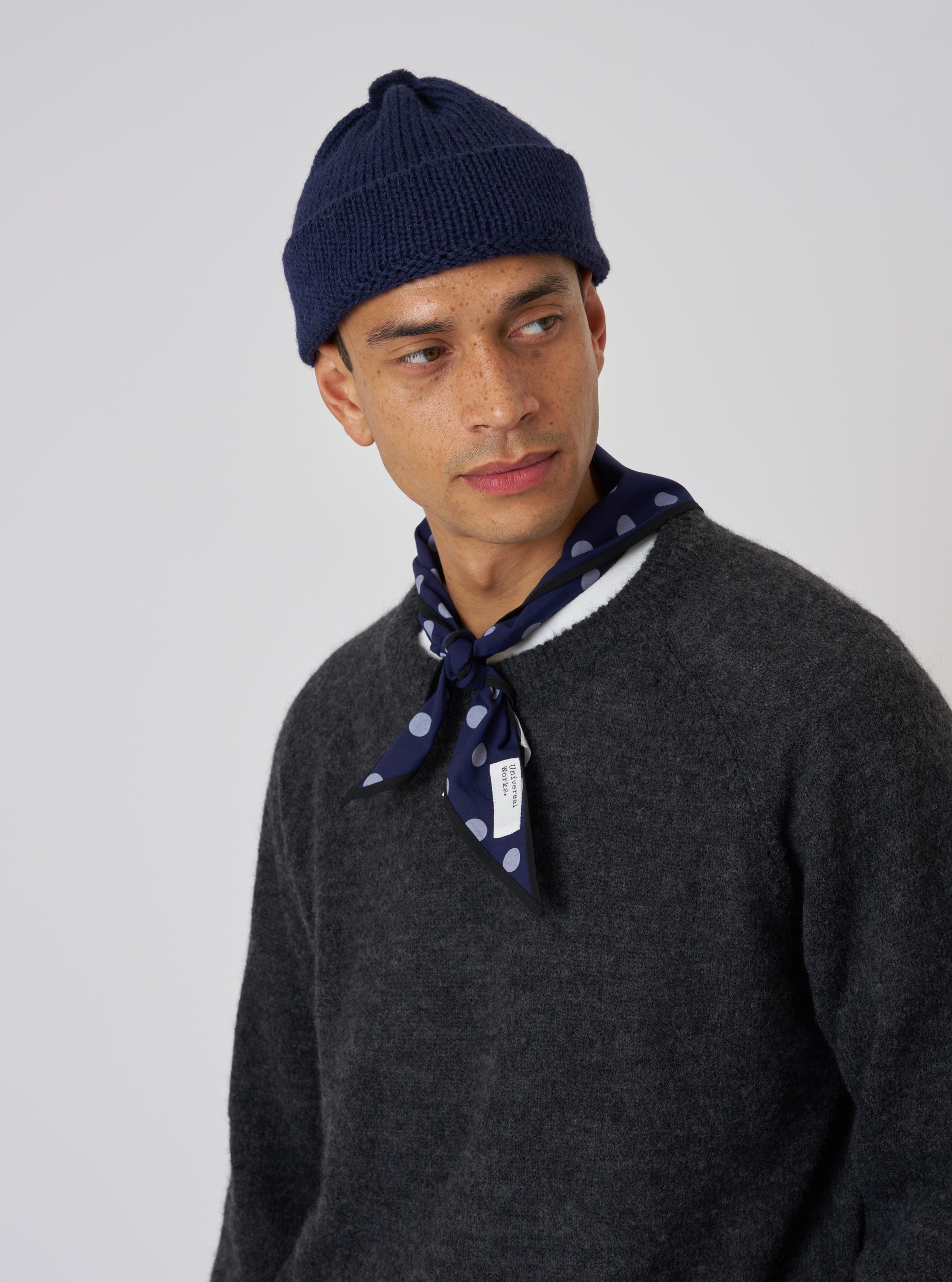 A model on a white background wears a Navy Bold Dot Print Rayon Scout Scarf tied around his neck with a gray sweater and navy beanie.
