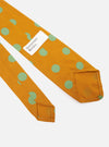 Close-up of a Mustard Bold Dot Print Rayon Tie on a white surface. The tie is mustard yellow with large green dots. A white label with black text is visible.