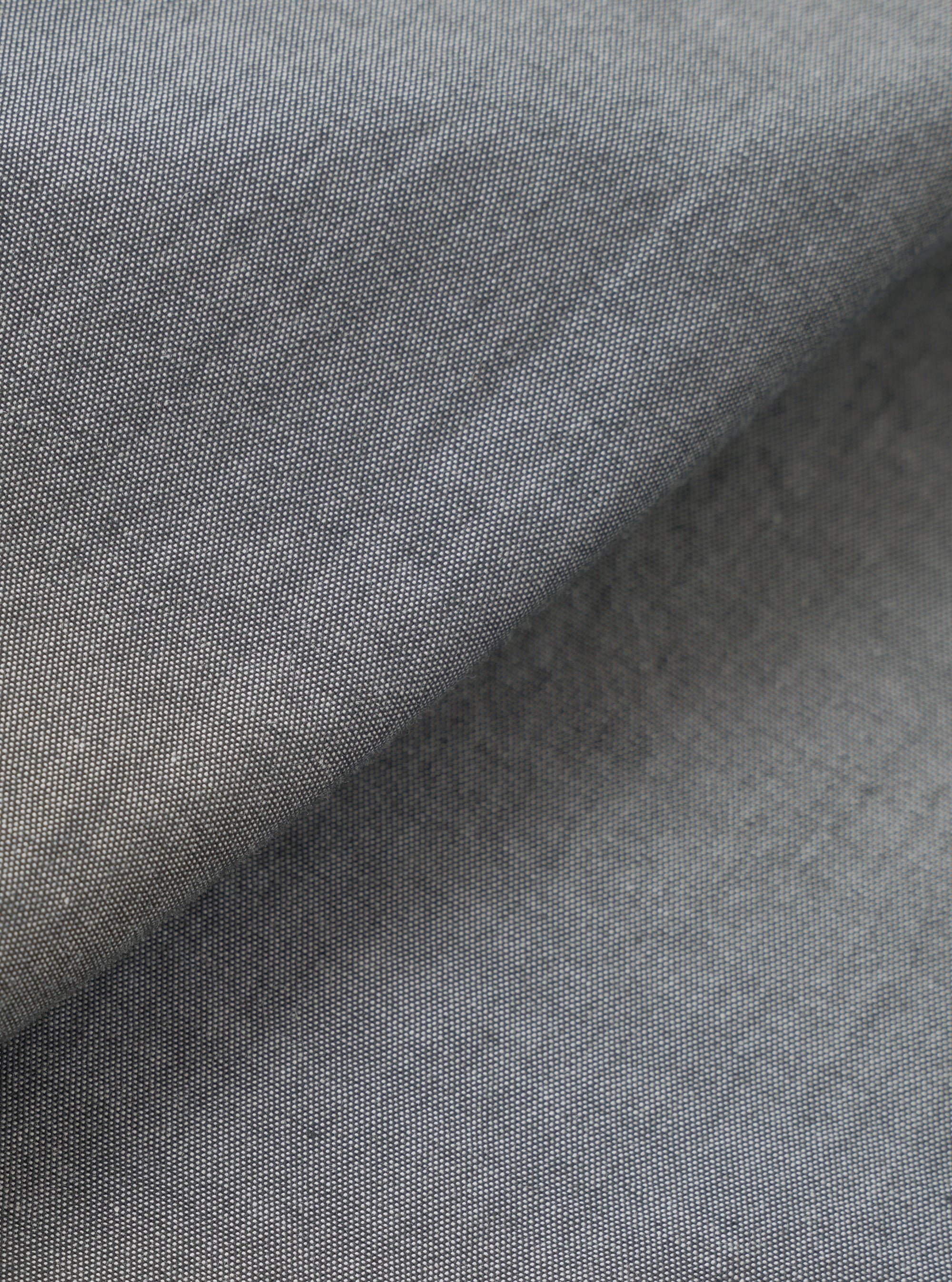 Close-up of the chambray fabric on the Black Prince Daybrook Shirt. The woven textile shows a fine two-tone grey and white yarn weave typical of chambray. A folded edge runs diagonally across the frame, revealing a soft brushed inner lining in a darker grey tone.