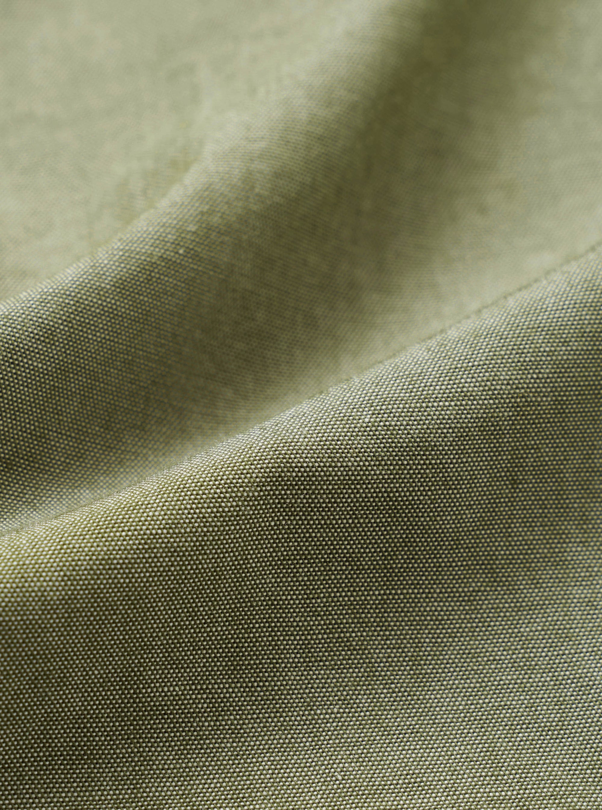 Close-up of the olive green chambray fabric of the Daybrook Shirt, showing a plain weave texture with a subtle two-tone yarn construction. A folded seam runs diagonally across the frame, highlighting the fabric weight and finish.