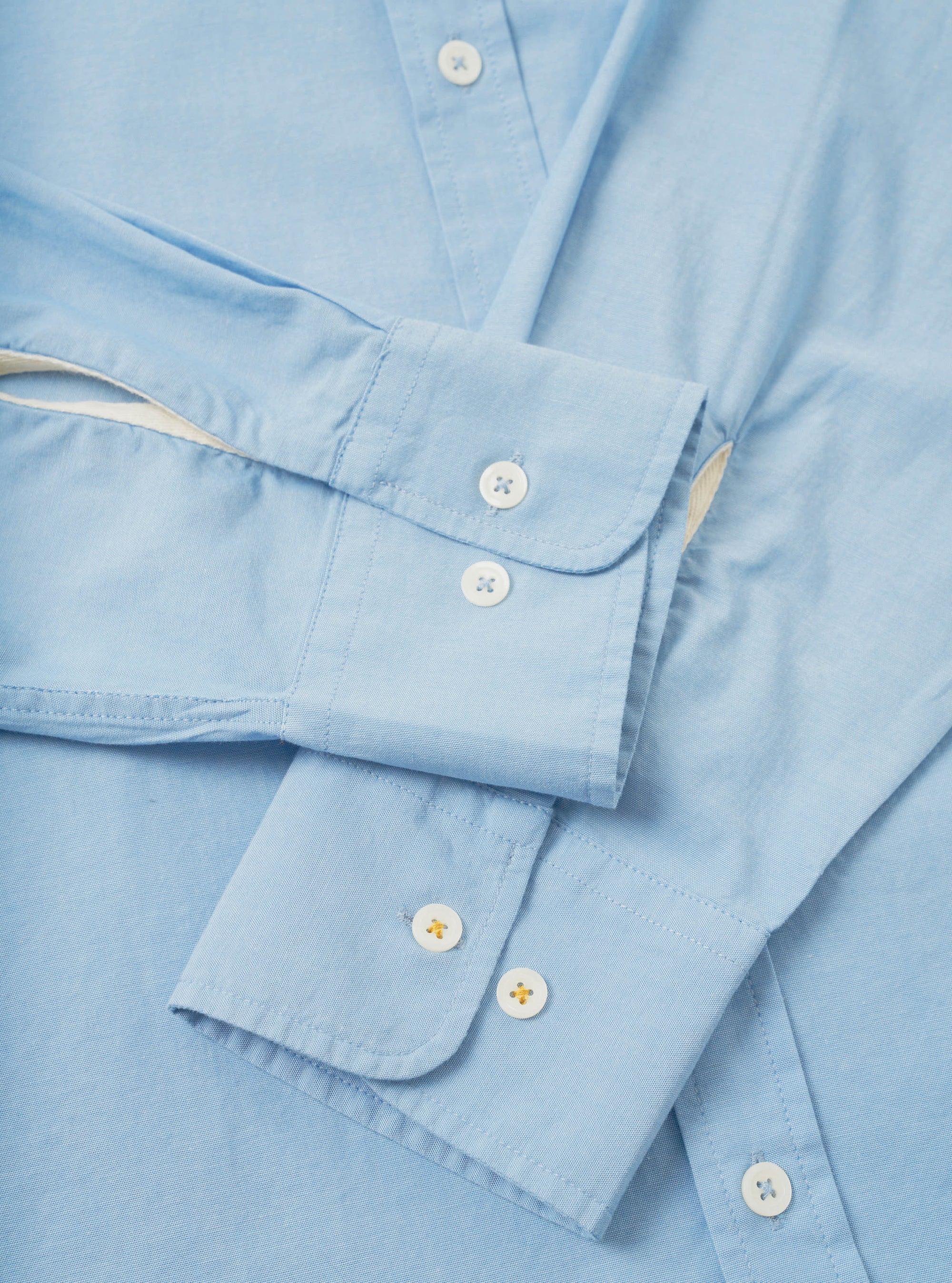 Close-up flat lay of the sky blue Daybrook shirt in chambray cotton, showing two buttoned barrel cuffs with rounded corners and white buttons with cross stitching. A cream garment tag is visible at the sleeve join. The placket with a single white button is visible at the top of the frame.