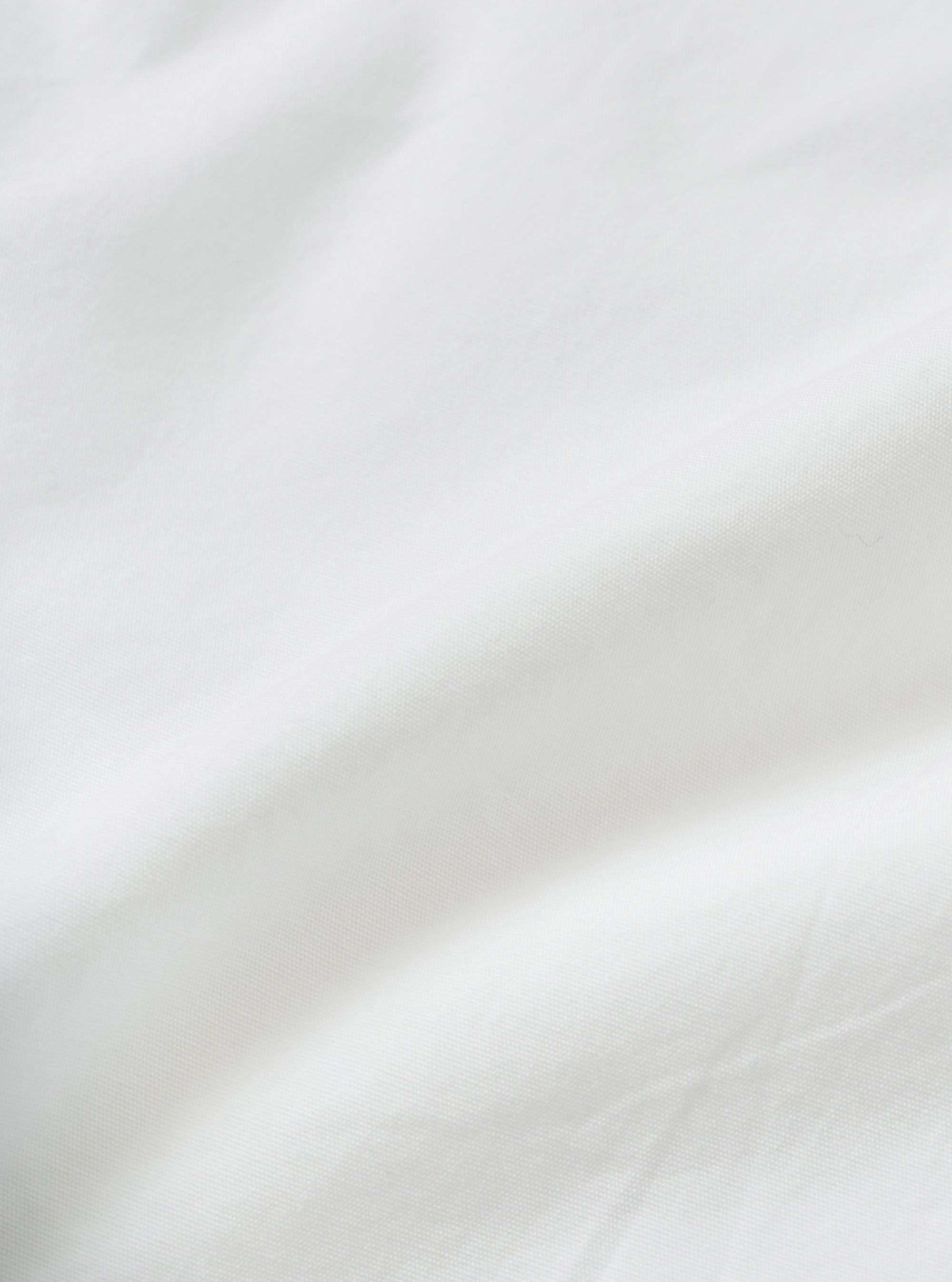 Close-up of the white chambray fabric used in the Daybrook shirt, showing a fine woven texture with a subtle grid-like weave structure. Soft natural folds run diagonally across the frame, highlighting the fabric's lightweight drape and clean white tone. No garment details or model visible.