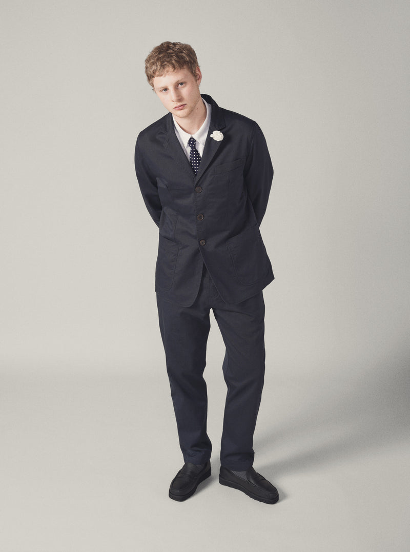 A model stands against a neutral backdrop wearing the Navy Twill London Jacket. The jacket is paired with a white shirt, polka dot tie, and black loafers for a smart, fitted look.