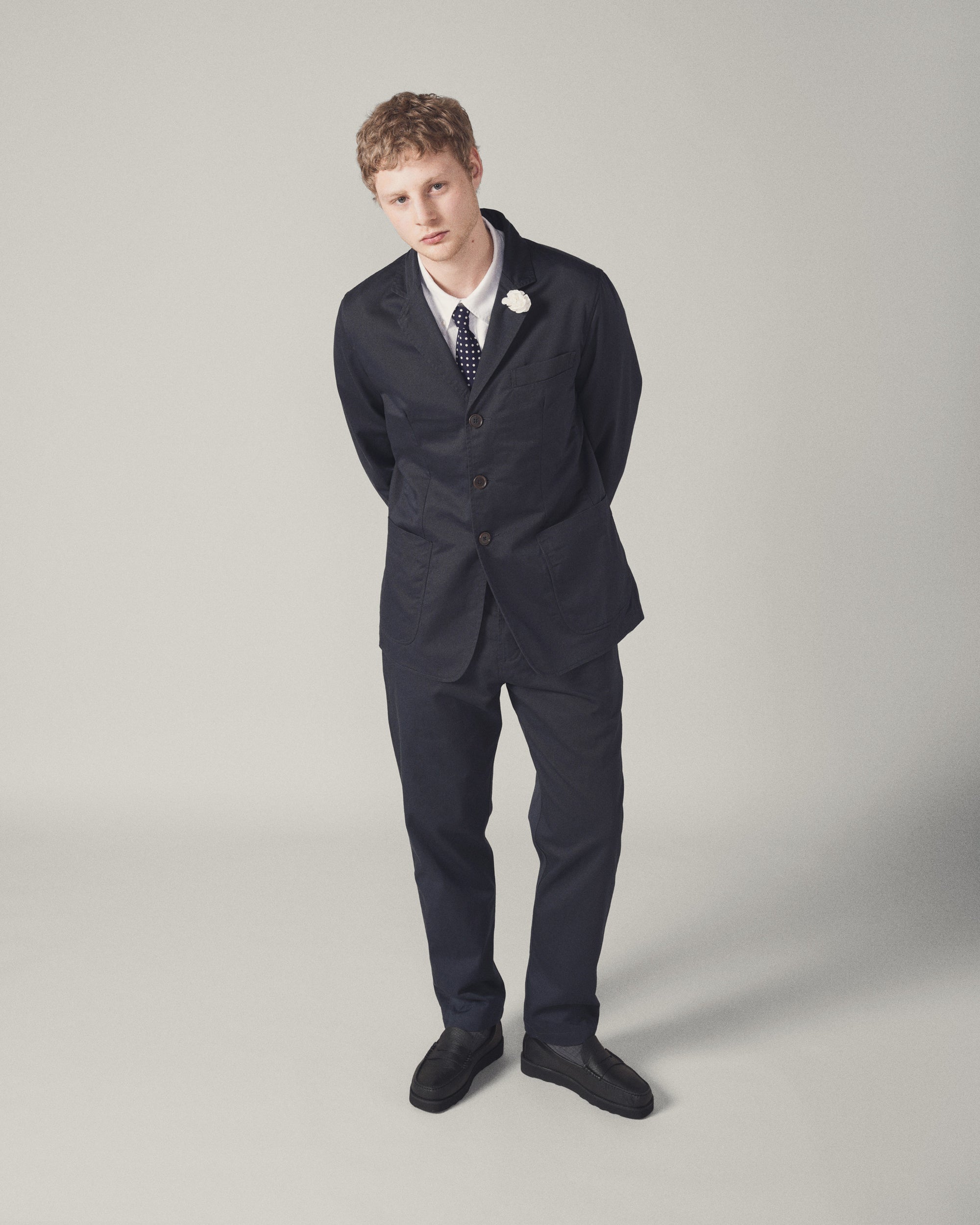 A model stands against a neutral backdrop wearing the Navy Twill London Jacket. The jacket is paired with a white shirt, polka dot tie, and black loafers for a smart, fitted look.