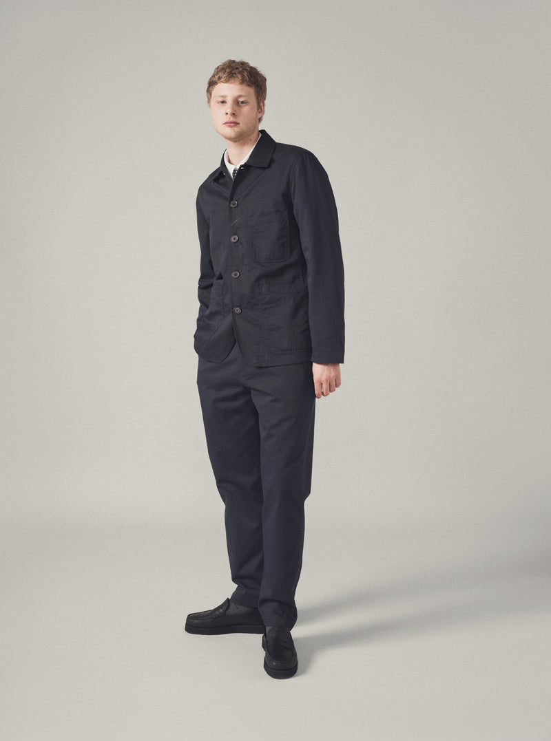 A model stands against a neutral backdrop wearing a Navy Twill Bakers Jacket and matching trousers. The jacket is buttoned and has a relaxed fit.