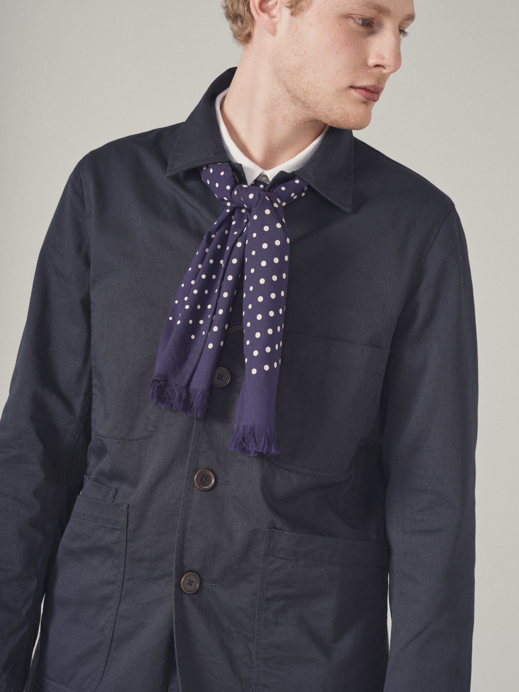 Close-up of a model wearing a Navy Twill Bakers Jacket with a polka dot scarf, against a neutral background. The jacket is a dark navy color and appears to be made of twill.