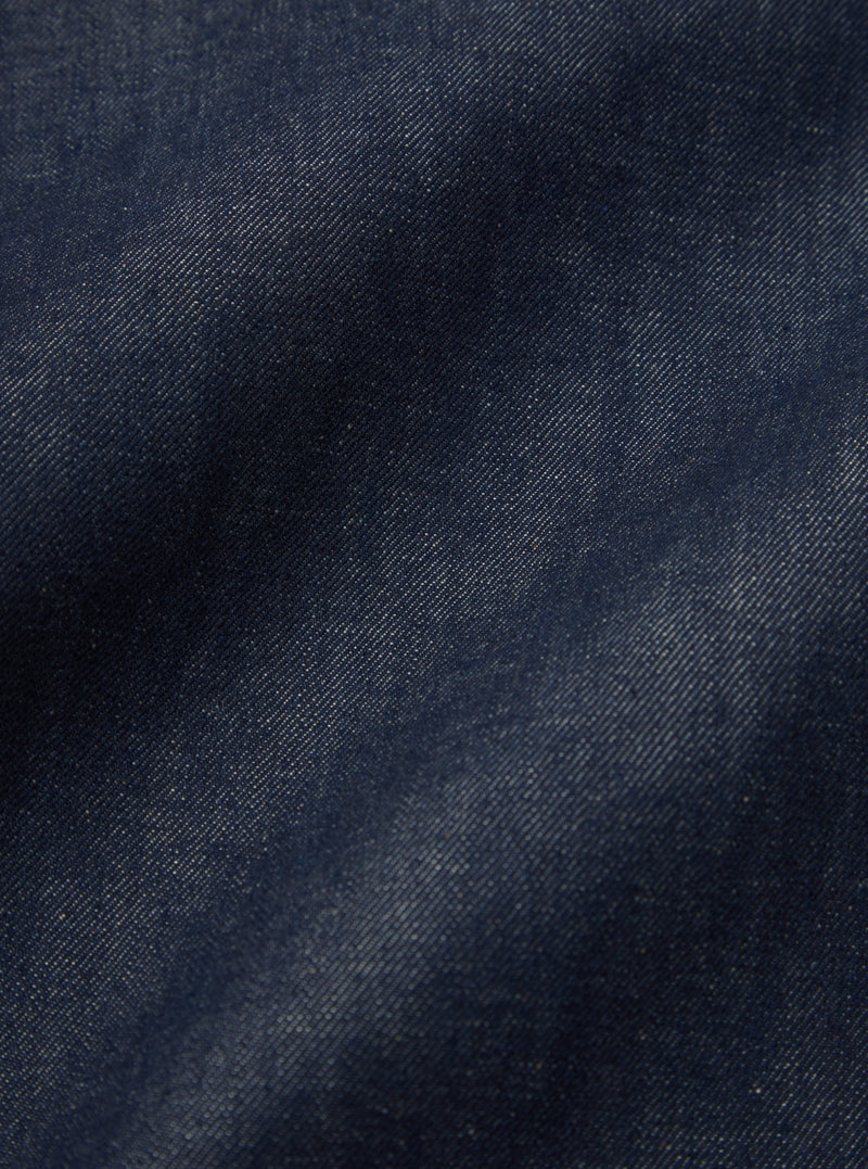 Close-up of the Indigo 13oz Selvedge Denim Bakers Jacket, showcasing the rich indigo color and textured selvedge denim material. The jacket is laid flat on a neutral surface.