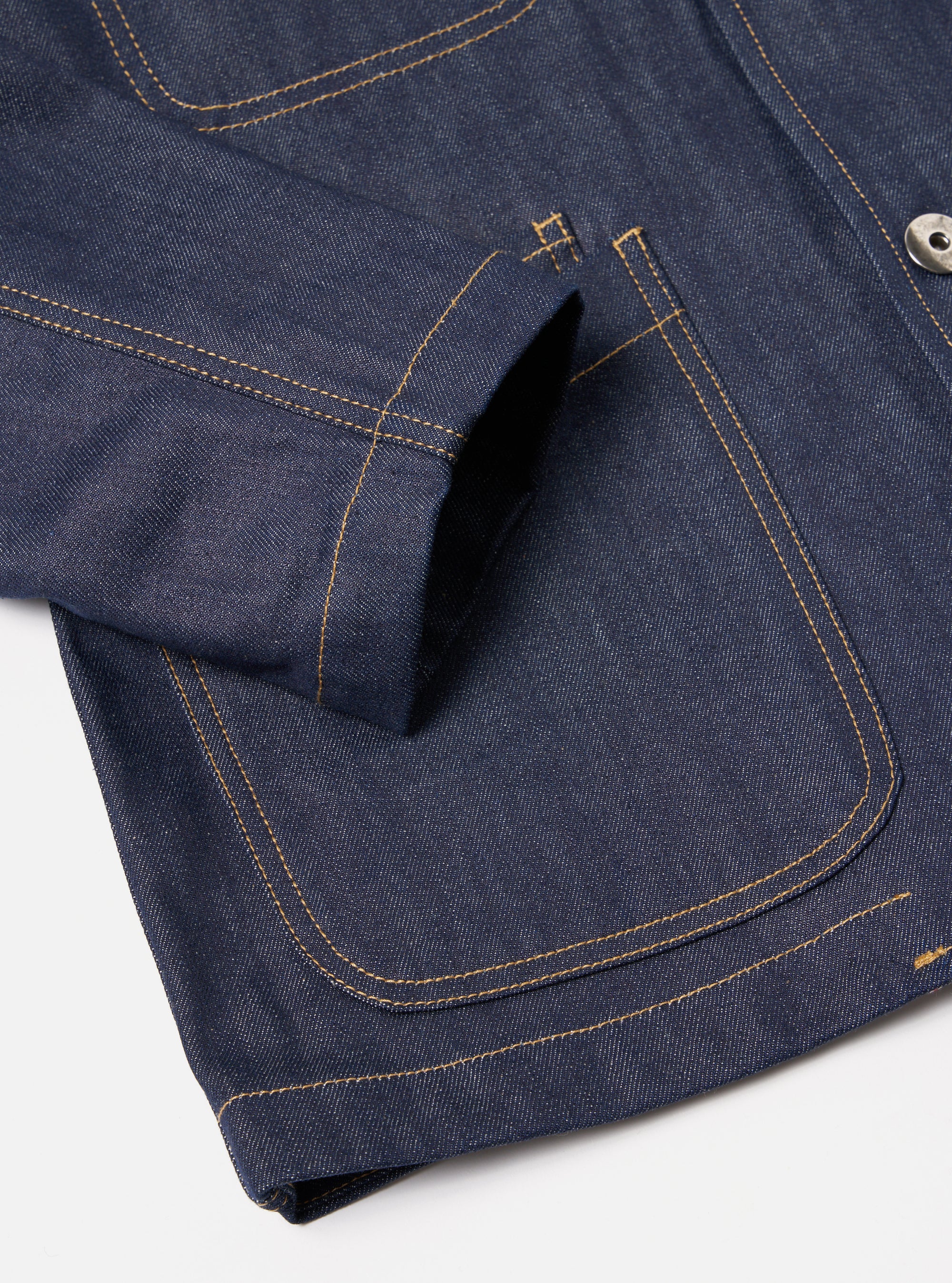 Close-up of the Indigo 13oz Selvedge Denim Bakers Jacket on a white surface. Shows the jacket's indigo denim, gold stitching, and pocket detail.