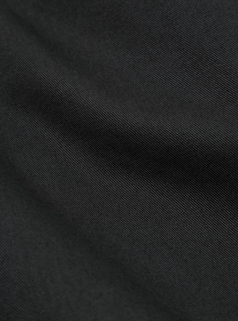 Close-up of the Black Twill Fatigue Pant fabric. The dark twill material is softly lit, showing the texture. The background is a simple, neutral surface.