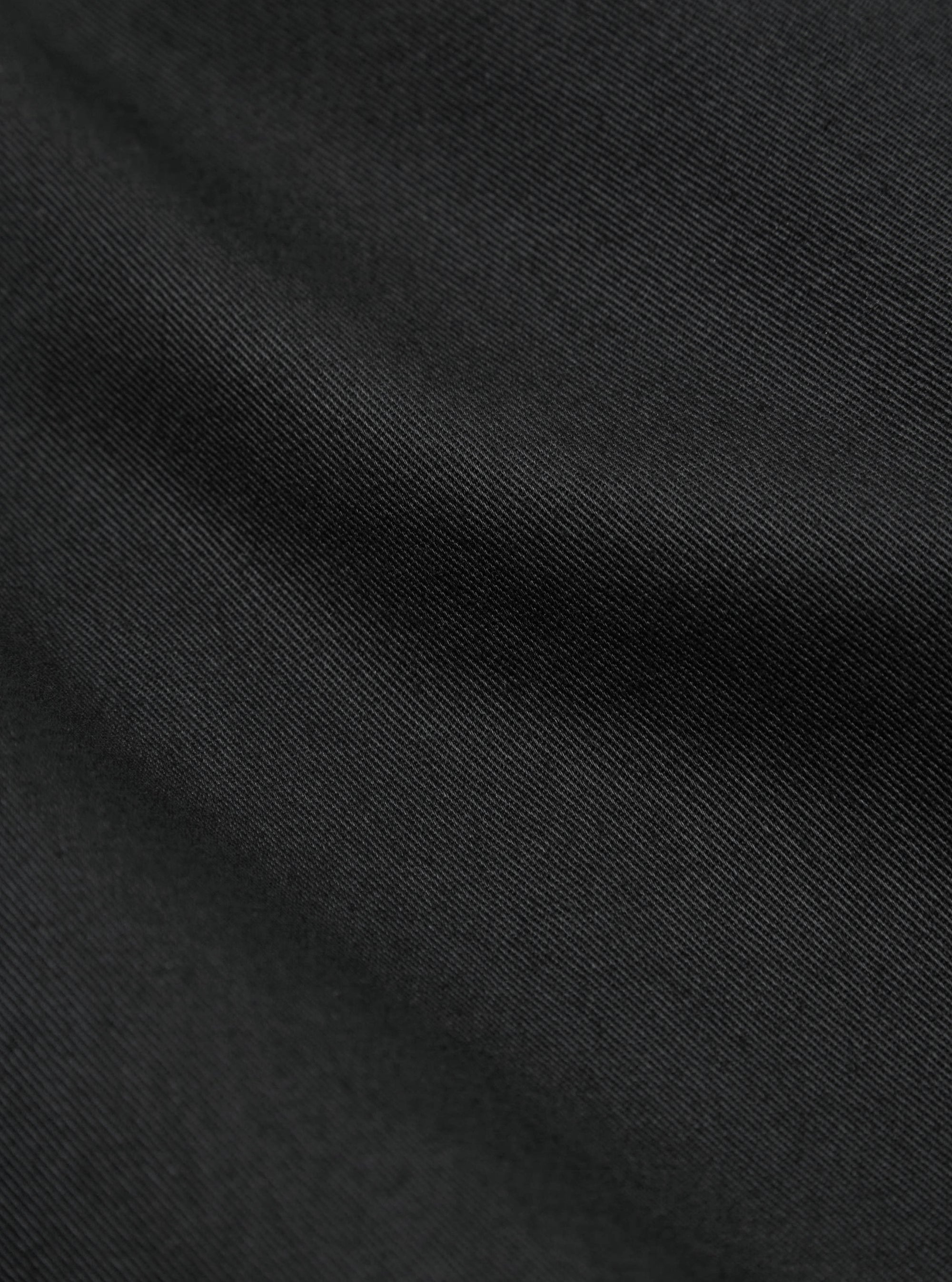 Close-up of the Black Twill Fatigue Pant fabric. The dark twill material is softly lit, showing the texture. The background is a simple, neutral surface.