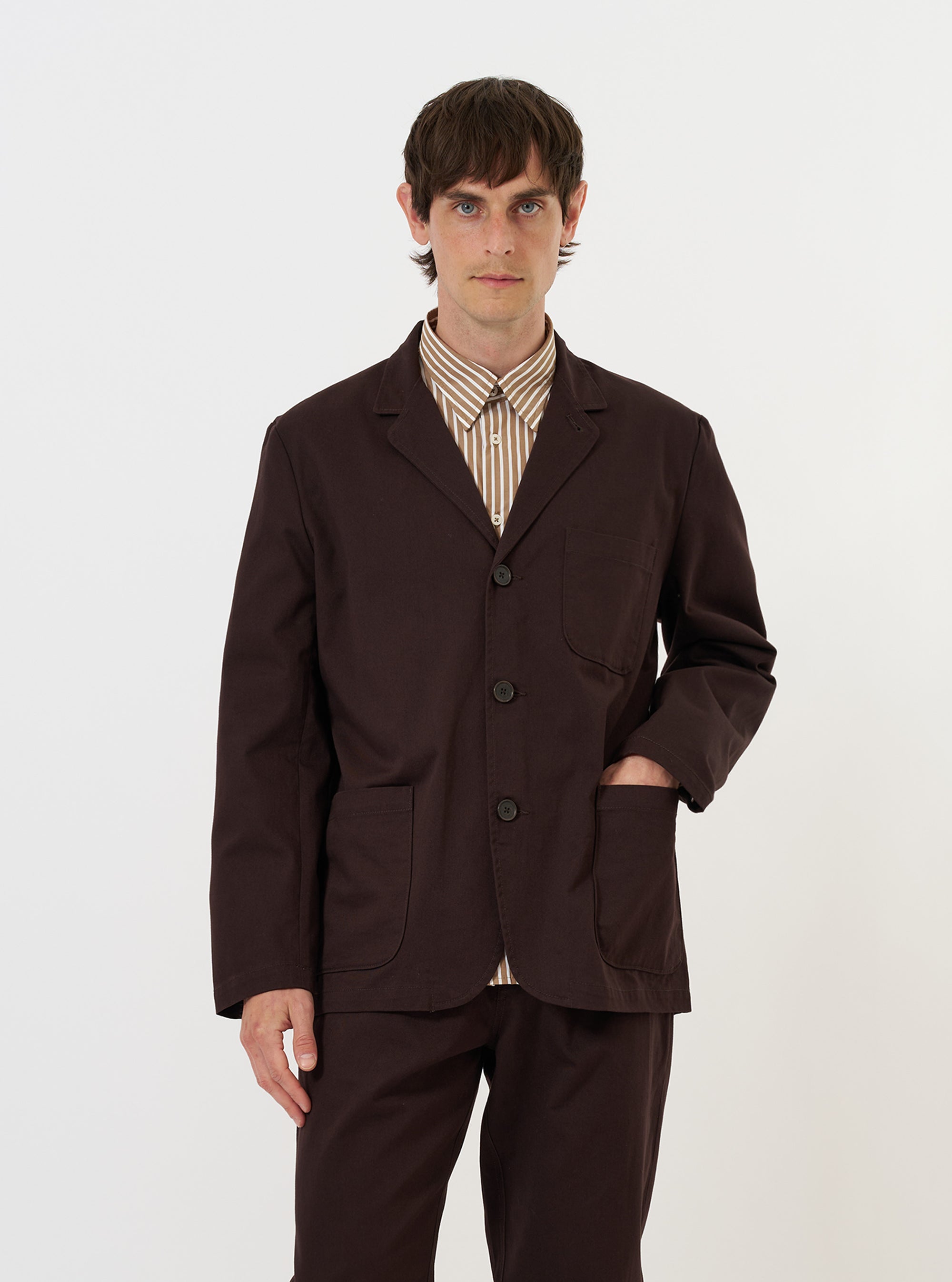 A male model stands against a plain white background wearing the Brown Twill Three Button Jacket. The jacket is a rich brown color with a textured twill weave, featuring a three-button front closure and three large patch pockets. It has a relaxed, unstructured fit with notched lapels and is paired with matching brown trousers for a complete tailored look.