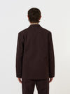 A model is wearing the Brown Twill Three Button Jacket against a plain white background. The image shows the jacket from the back, highlighting the relaxed fit, unstructured shoulders, and two-piece sleeves. The model is also wearing white trousers.