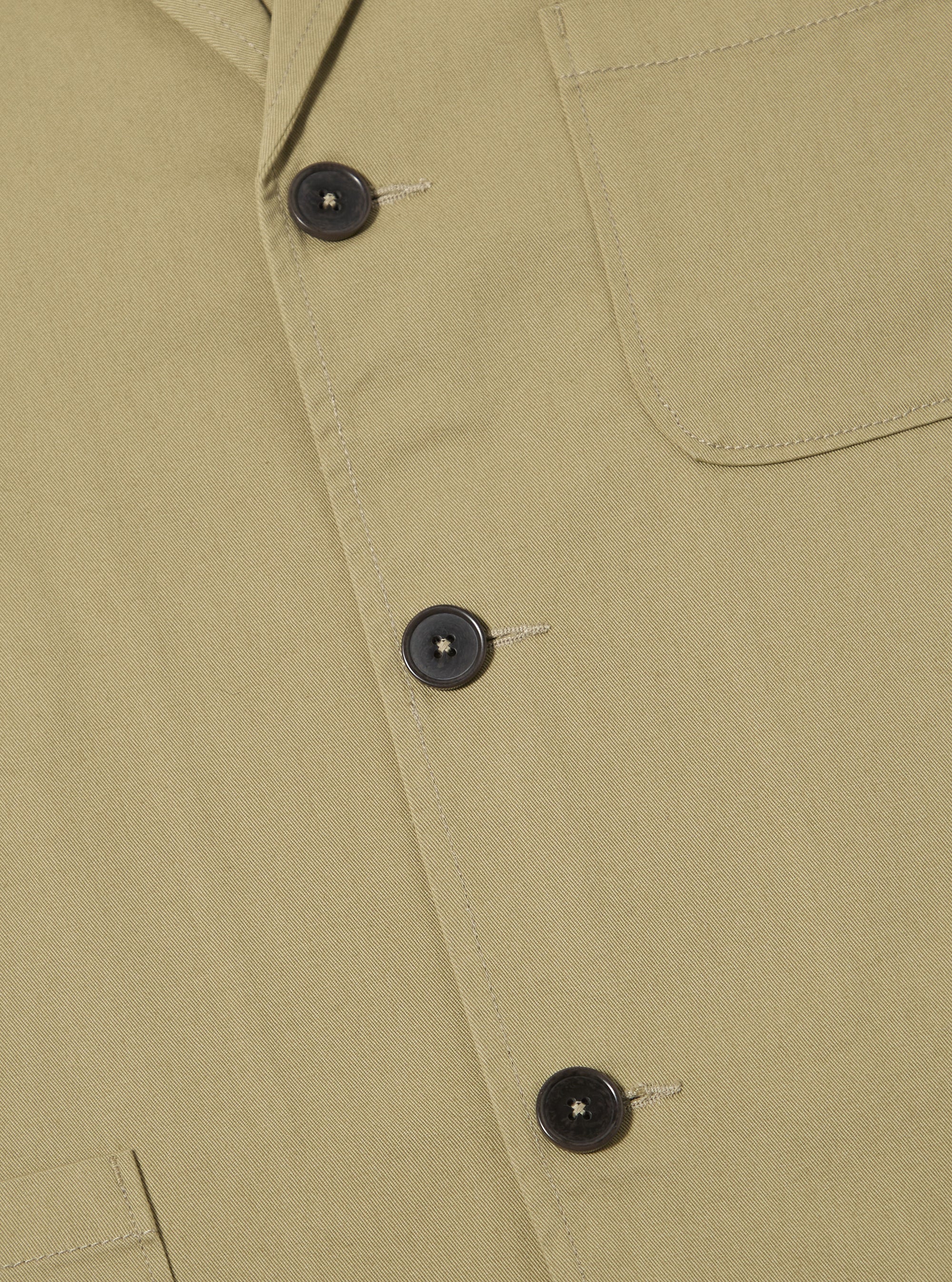 Close-up of the sand twill three button jacket on a flat surface. The jacket is a light tan color with dark brown buttons. The background is a plain white.