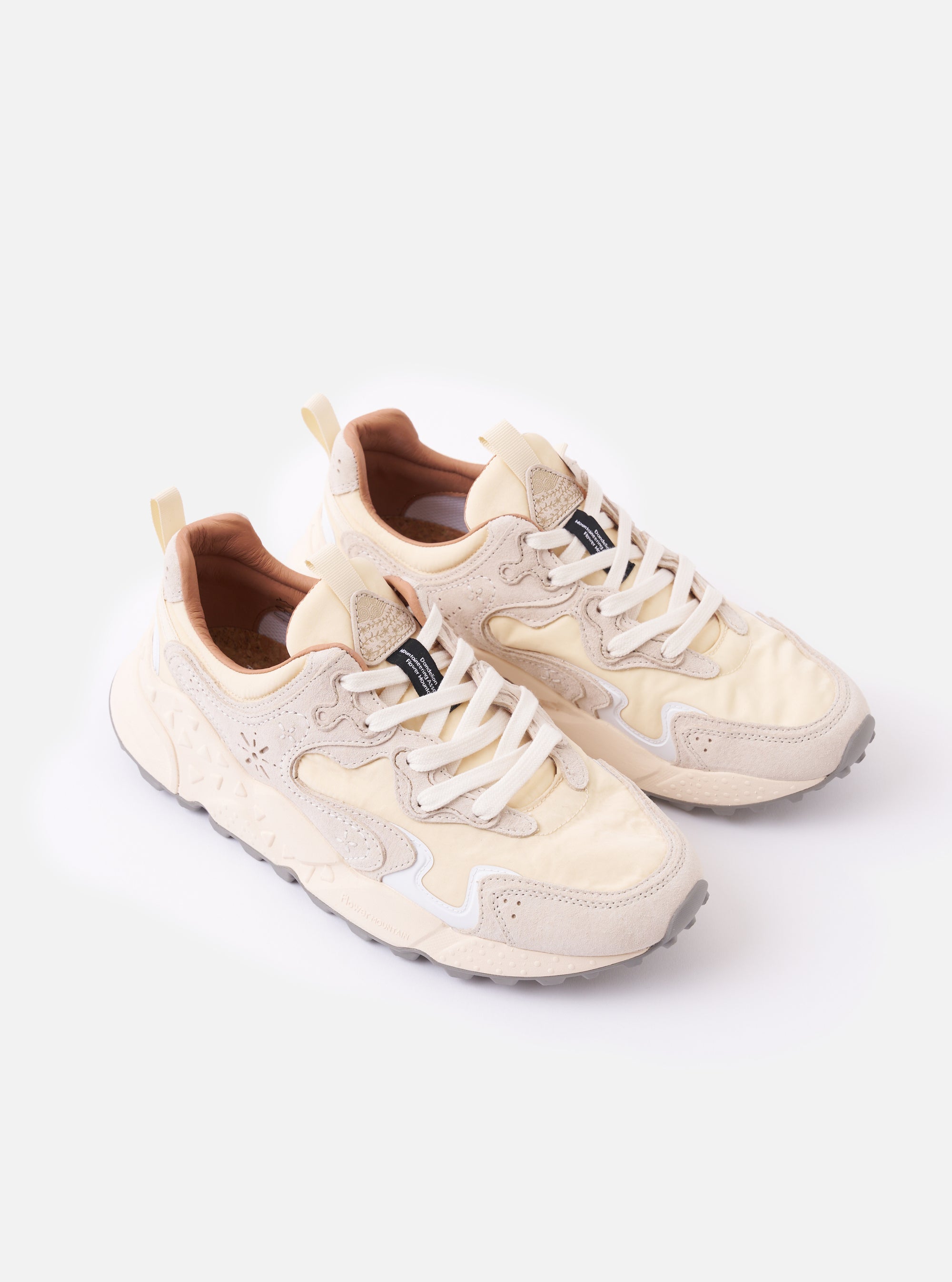 The Flower Mountain Milk/Light Beige Suede/Nylon Rulis Uni sneaker is shown in a detail view as a flat lay against a plain white background. The image highlights the layered construction of the footwear, featuring overlapping light beige suede panels and milky white technical fabric. The thick, moulded rubber sole with a textured sidewall and the webbing pull tab at the heel are prominent.