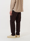 A model wears the Brown Twill Military Chino against a plain white background. The image shows the model from the waist down, highlighting the slim, straight leg fit of the brown cotton trousers. The model is standing with one leg slightly forward, showcasing the fabric's texture and the garment's clean silhouette.