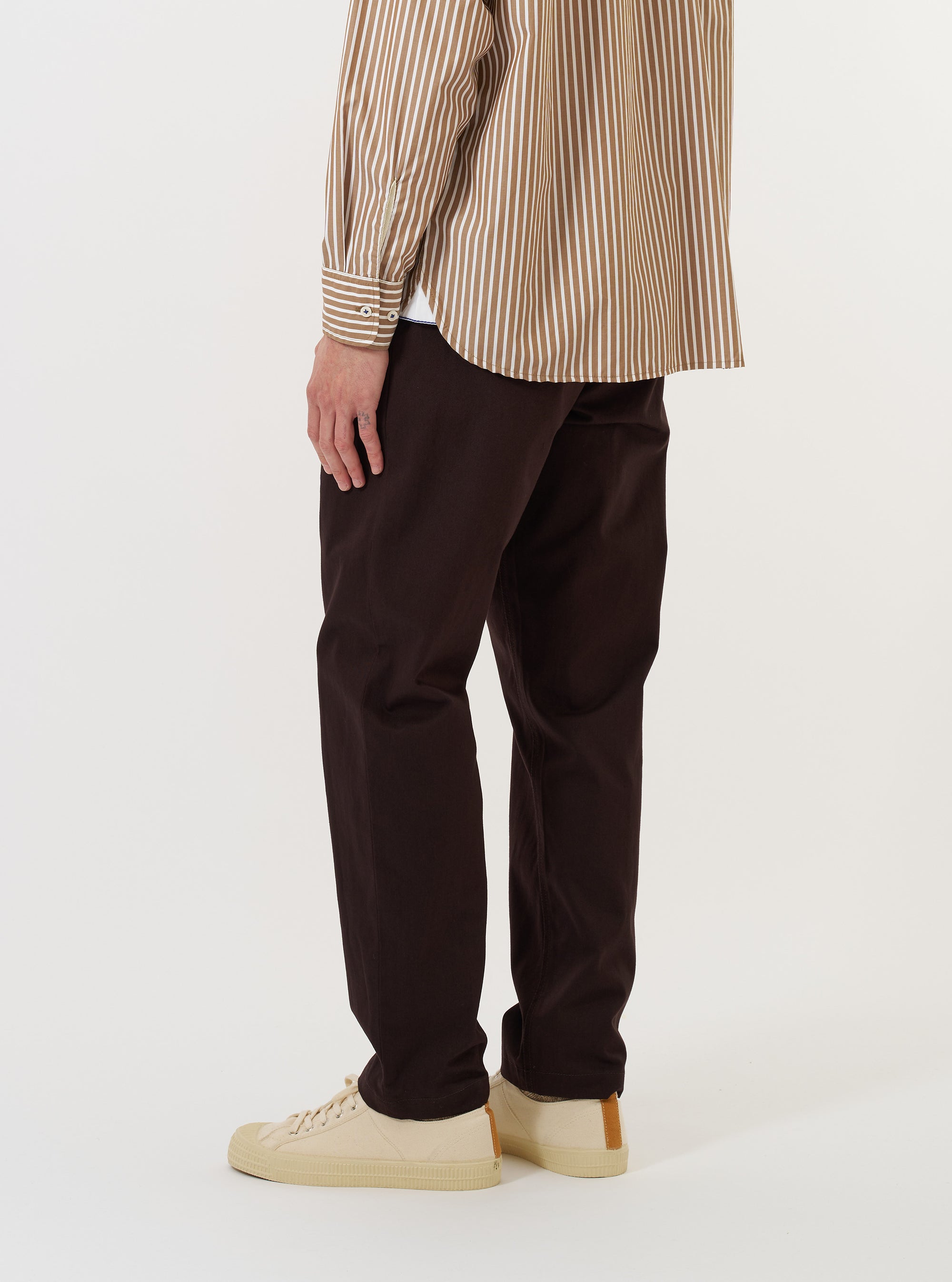 A model wears the Brown Twill Military Chino against a plain white background. The image shows the model from the waist down, highlighting the slim, straight leg fit of the brown cotton trousers. The model is standing with one leg slightly forward, showcasing the fabric's texture and the garment's clean silhouette.