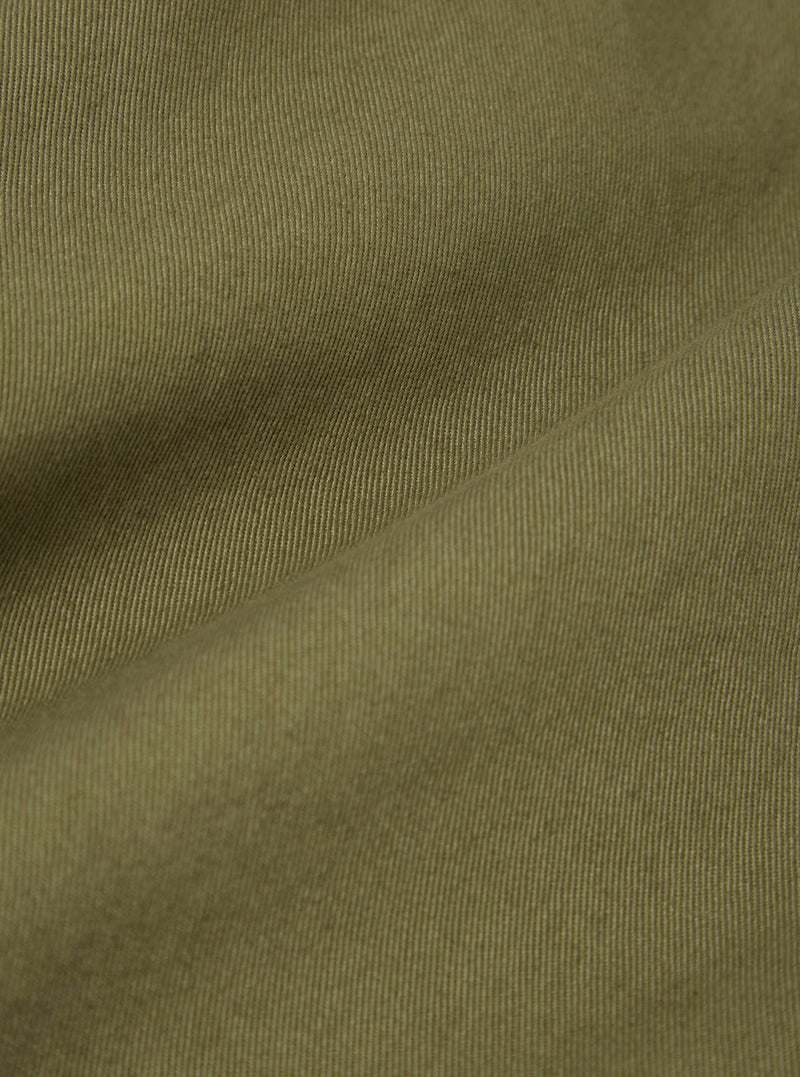 Close-up of the light olive twill fabric of a Baker's Jacket, showing the texture and color. The background is a plain surface.