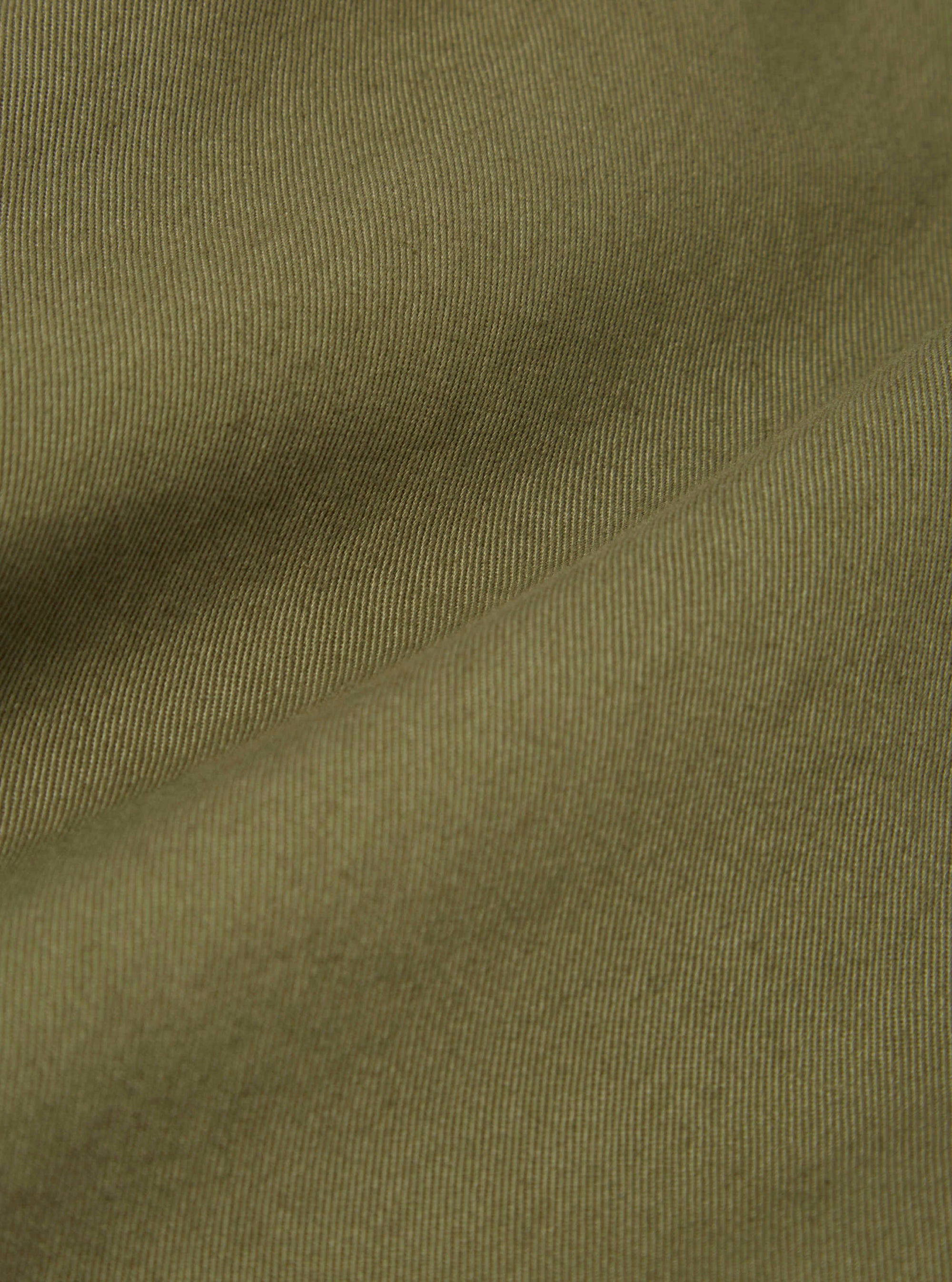 Close-up of the light olive twill fabric of a Baker's Jacket, showing the texture and color. The background is a plain surface.