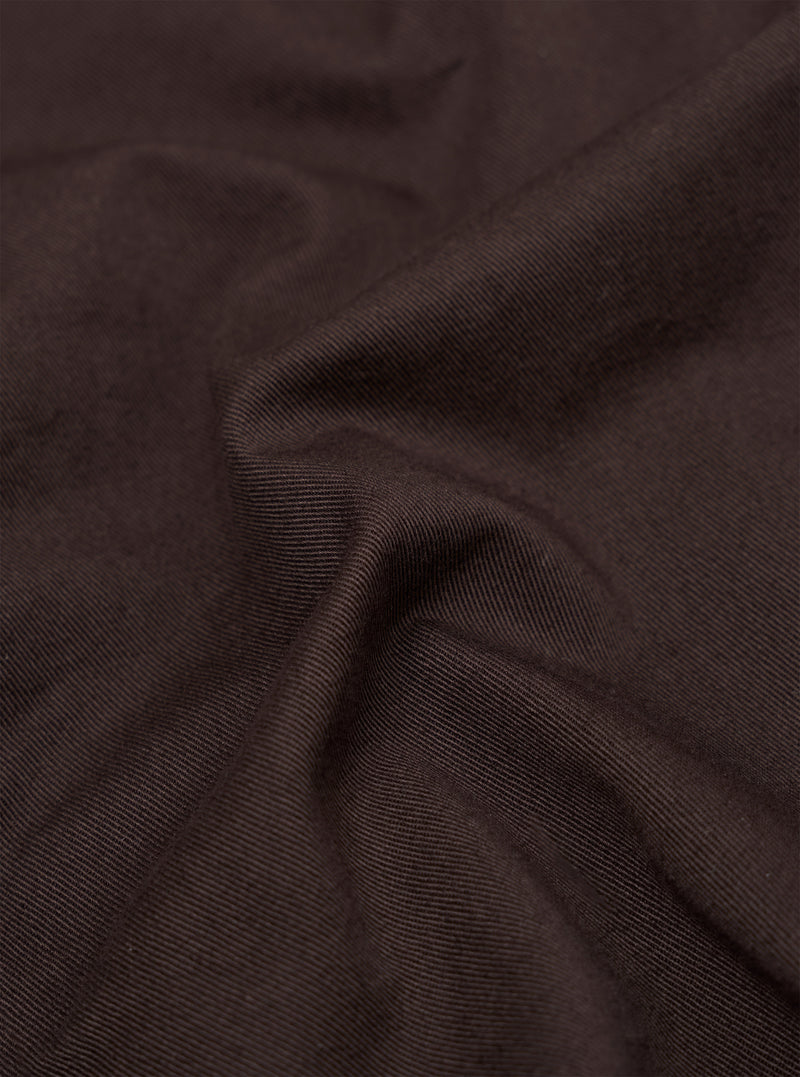 A close-up fabric swatch of the Brown Twill Pleated Track Pant shows a detailed view of the brown cotton twill material. The image highlights the diagonal weave and textured surface of the fabric.