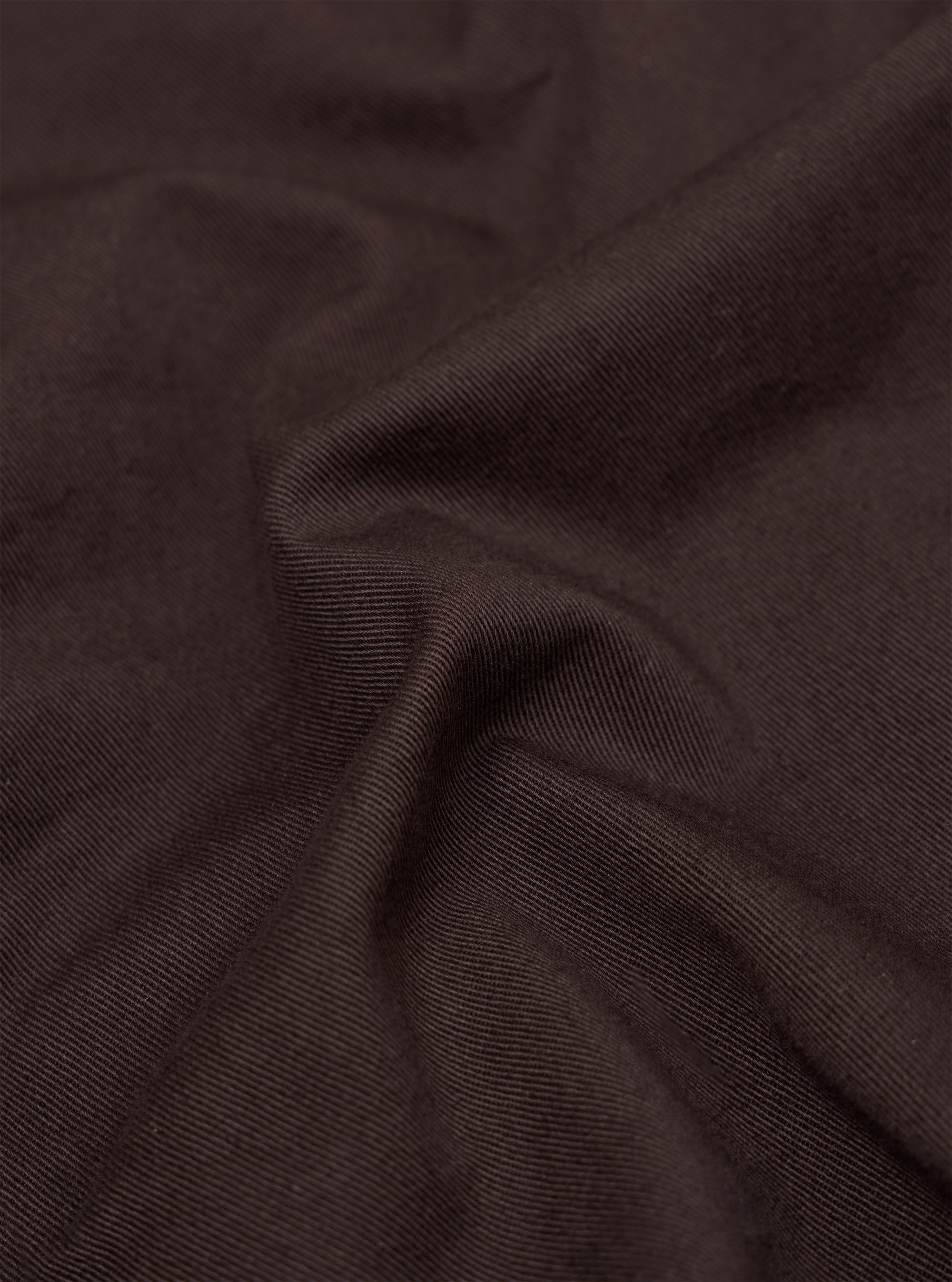 A close-up fabric swatch of the Brown Twill Pleated Track Pant shows a detailed view of the brown cotton twill material. The image highlights the diagonal weave and textured surface of the fabric.