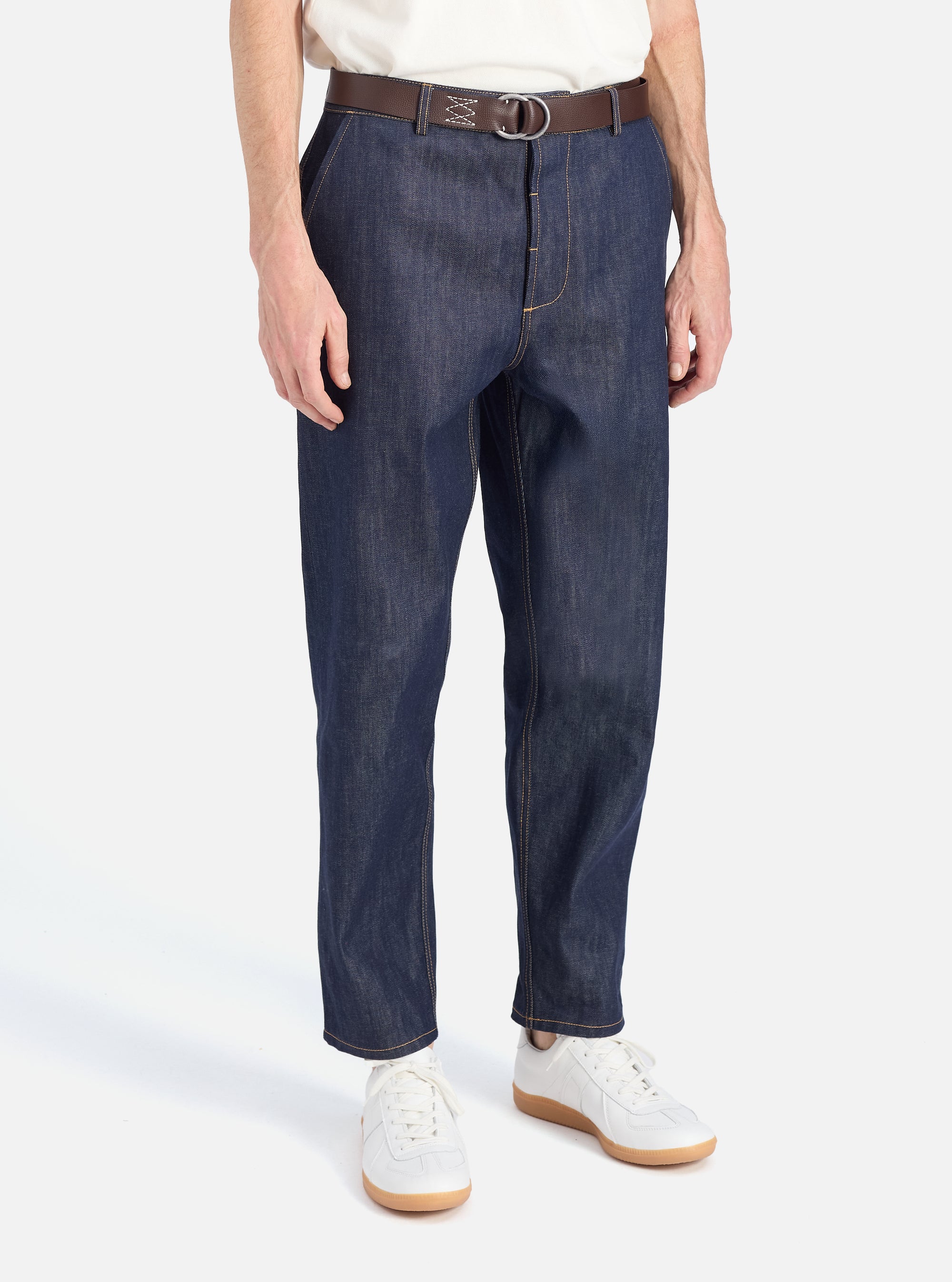 A model wears Indigo 13oz Selvedge Denim Military Chino, styled with a brown belt and white sneakers. Shot in a bright studio, the denim's texture and fit are highlighted.