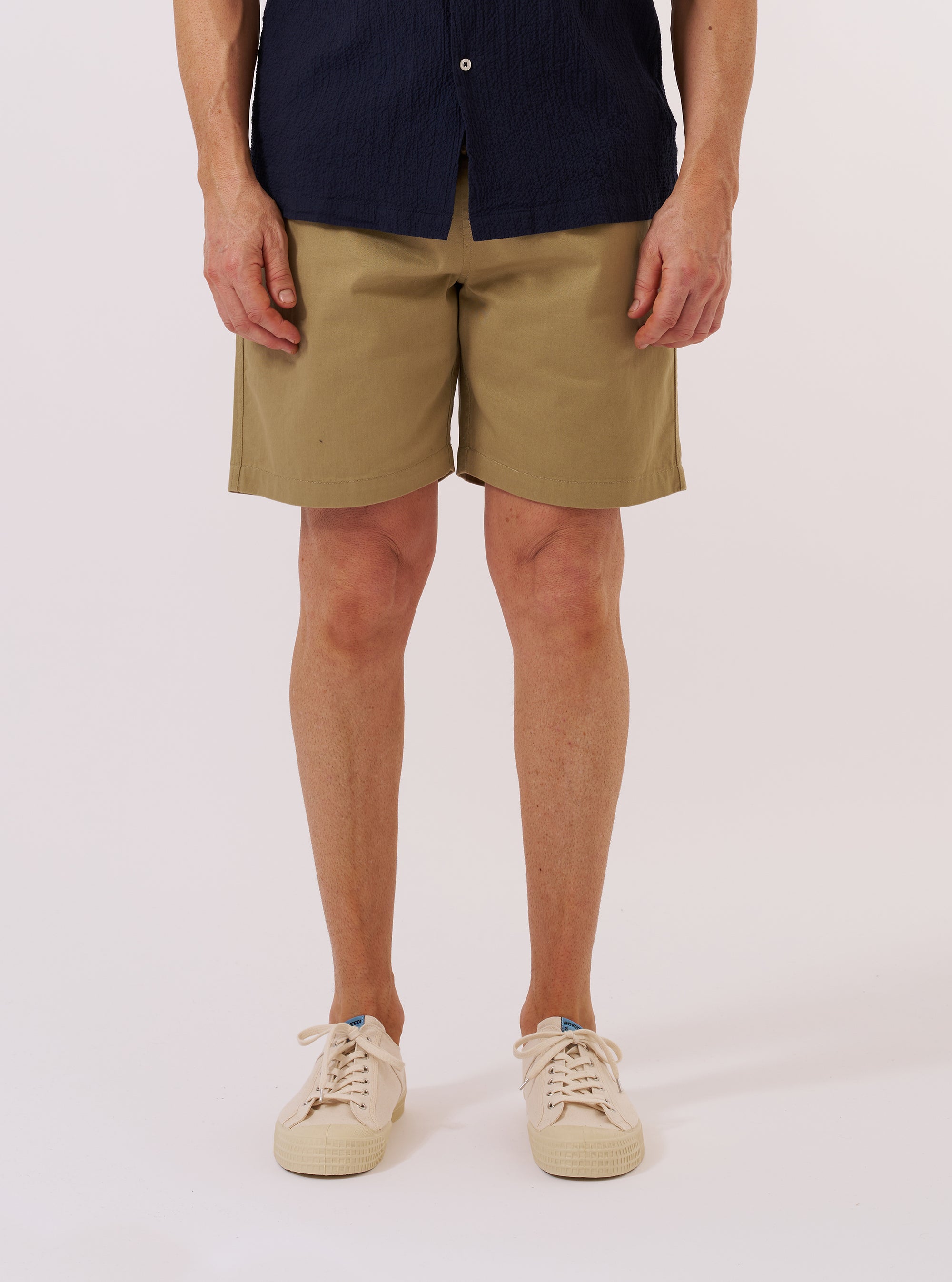 The Sand Twill Beach Short is worn by a model against a plain white background. The image shows a front view of the shorts, highlighting the elasticated waistband with a drawstring, two side pockets, and the sand-colored twill fabric. The model is wearing a white T-shirt tucked into the shorts to make the waistband prominent.