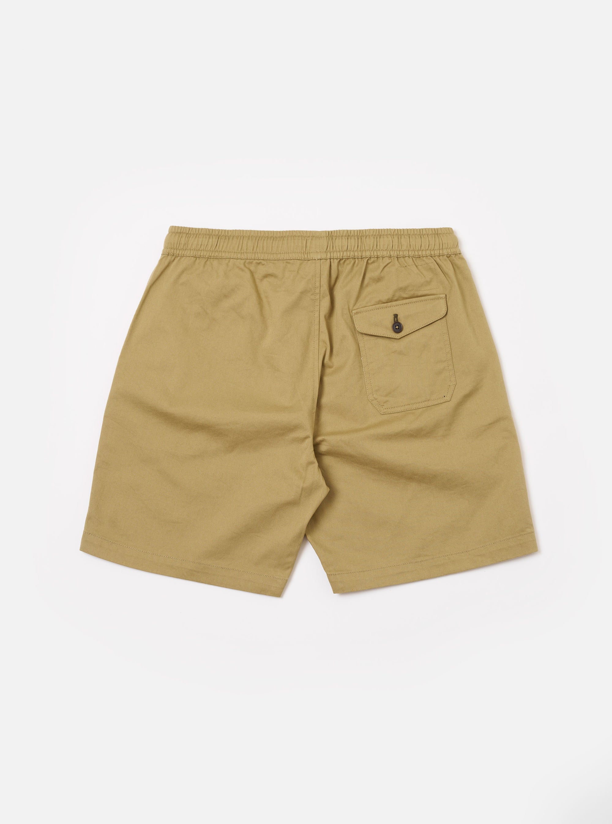 The Sand Twill Beach Short is presented as a flat lay from the rear against a plain white background. The shorts are a light sand color and feature a visible elasticated waistband and a single square patch pocket on the right side. The cotton twill fabric has a fine diagonal weave.