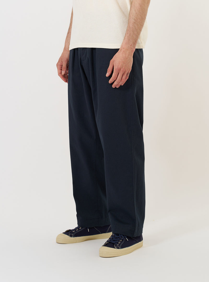 A model is wearing the Navy Twill Oxford Pant against a plain white background. The navy blue trousers feature a relaxed fit with a wide leg, a single pleat at the front, and a drawstring at the elasticated waistband. The model is also wearing a light-colored shirt and white sneakers.